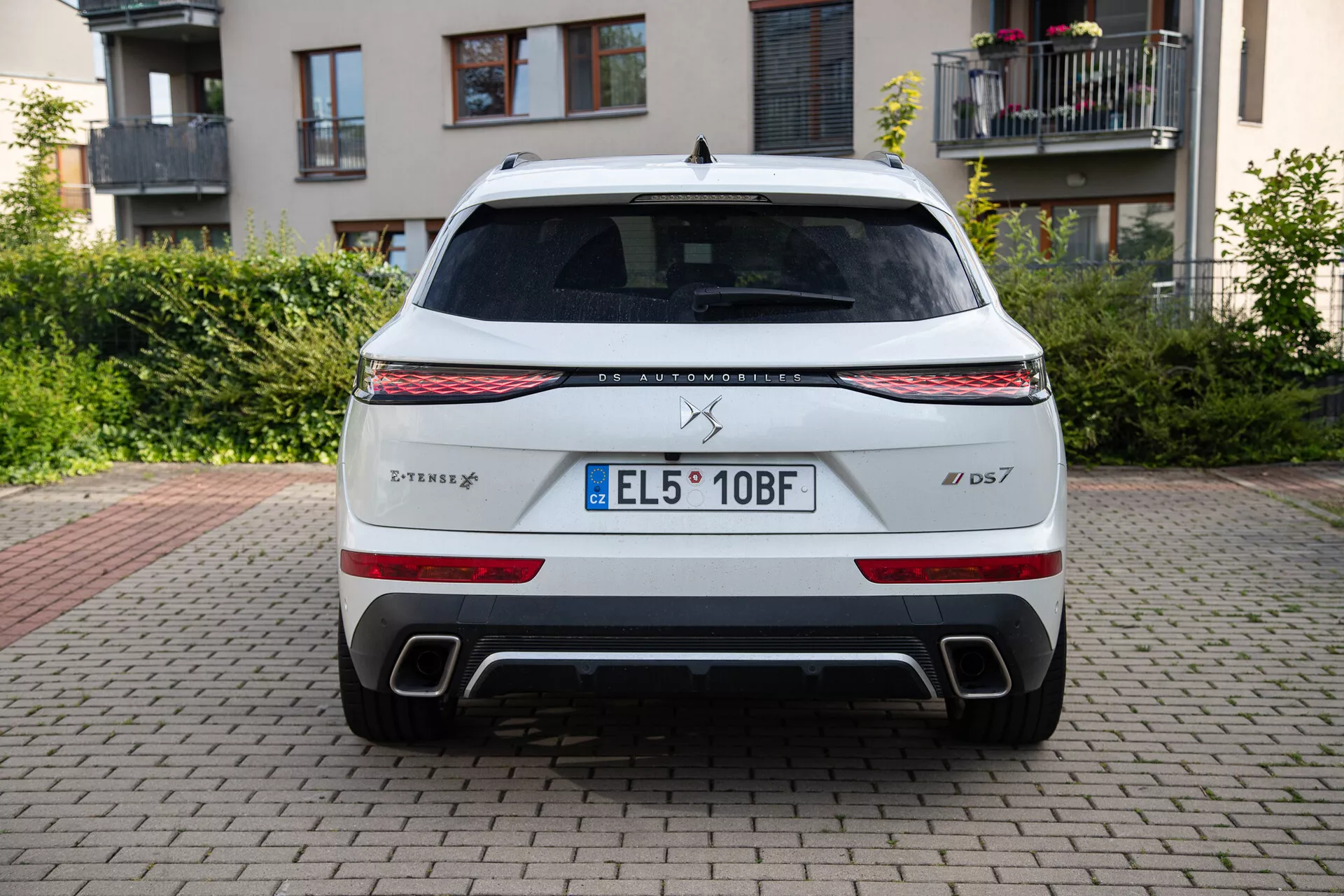 Vista trasera de un DS Automobiles DS 7 2022 estacionado en superficie de ladrillo con edificios y vegetación al fondo, mostrando tira de luz LED, luces traseras, salidas de escape dobles y placa de matrícula de la UE