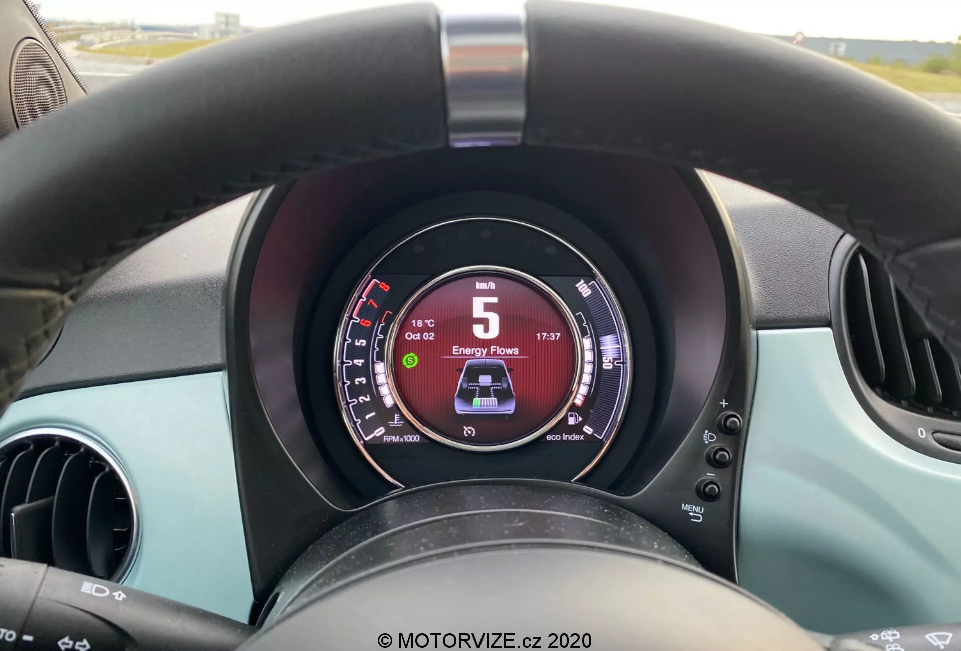Vista de cerca desde la perspectiva del conductor del interior de un Fiat 500 2015, destacando un volante negro ligeramente desgastado, un panel de instrumentos digital que muestra 5 km/h y 'D' para conducir, dos ventilaciones circulares y acentos de color turquesa en el tablero y las puertas.