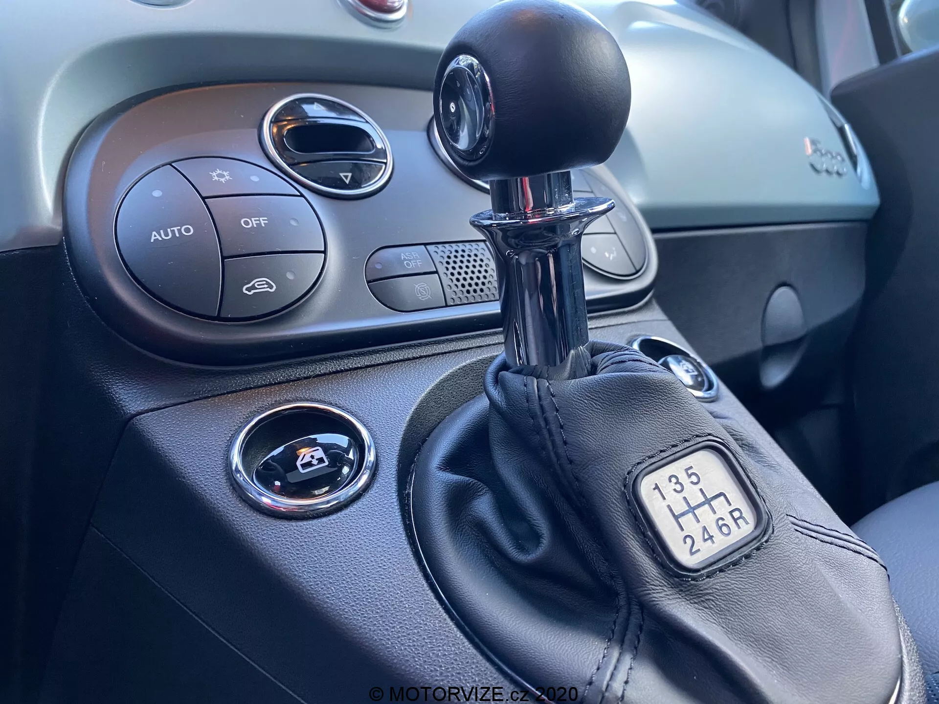 Vista de cerca de la consola central dentro de un vehículo Fiat 500 2015, centrada en el lado derecho. La imagen muestra una palanca de cambios manual con configuración de 6 velocidades, parte de la consola central con controles de clima etiquetados como 'AUTO', botón 'OFF', botón de arranque/parada para el encendido, ventilación circular y el salpicadero texturizado.