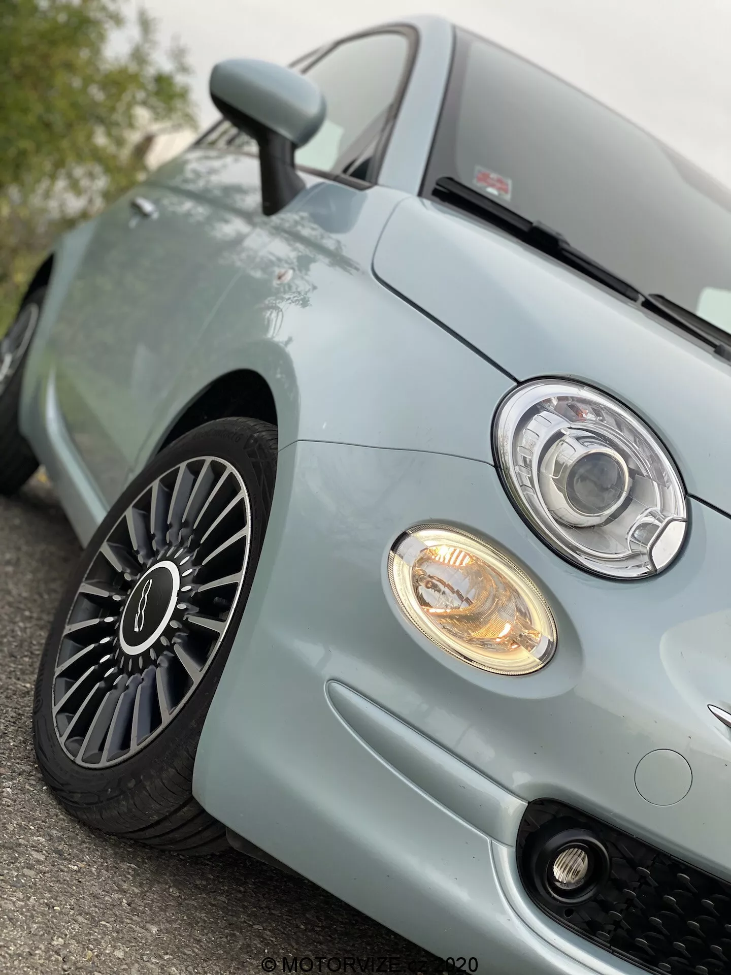 Vista de primer plano en ángulo bajo del cuarto delantero izquierdo de un Fiat 500 2015 azul claro, que muestra el faro de estilo proyector moderno con un anillo LED o halo, luz antiniebla iluminada, rejilla deportiva de malla de panal, sección delantera del capó y llanta de aleación de múltiples radios con buena profundidad de la banda de rodadura, sugiriendo un vehículo bien mantenido y avanzado diseño automotriz.