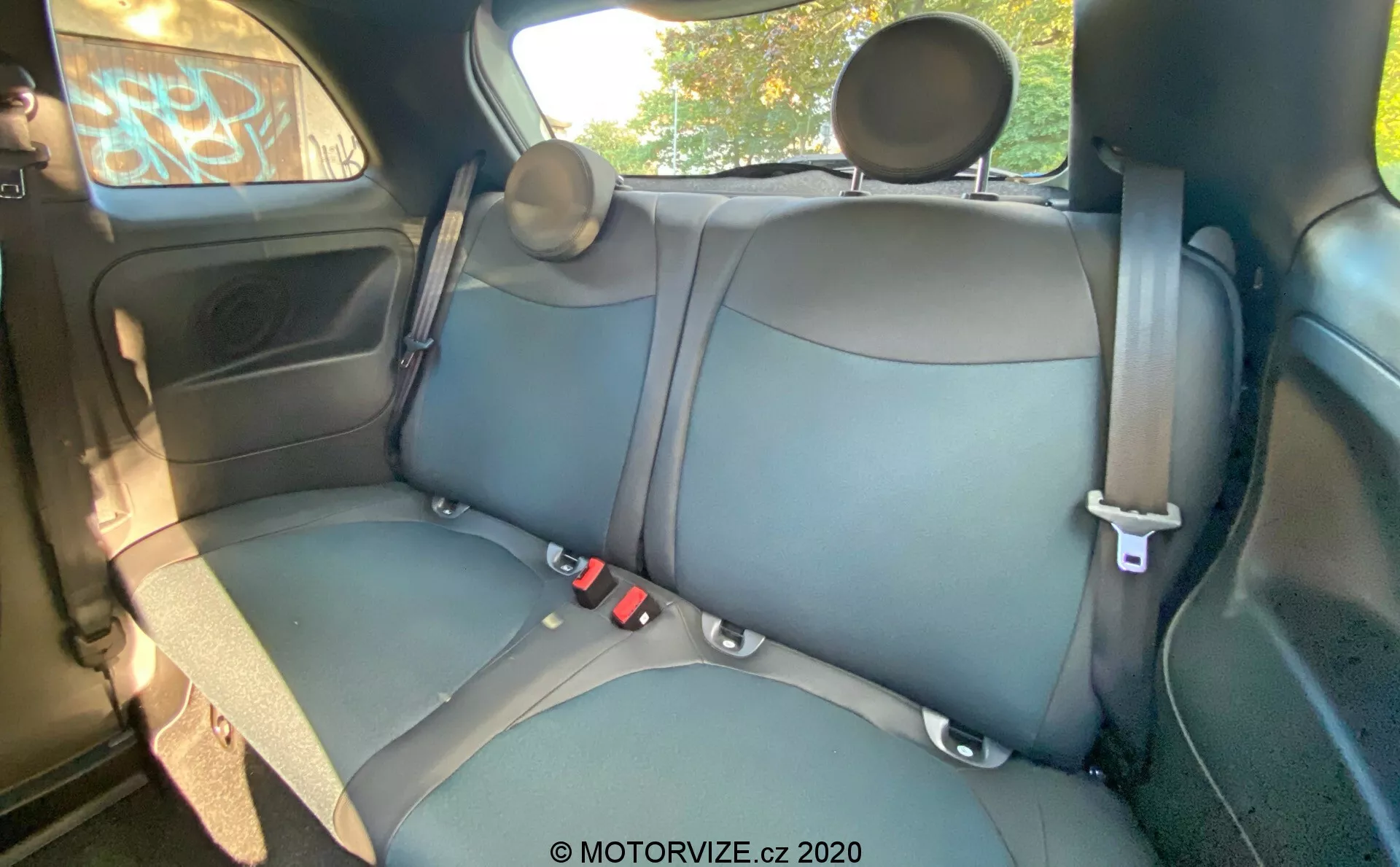 Interior de un Fiat 500 de 2015 que muestra los asientos traseros con tapicería de cuero o cuerina en color oscuro, costuras visibles, cinturones de seguridad de tres puntos, reposacabezas ajustables, panel de la puerta con control de ventana eléctrica, compartimento de almacenamiento, rejilla de altavoz, vista parcial del techo y asa superior. El ángulo de la cámara está ligeramente elevado de delante hacia atrás, proporcionando una vista detallada del asiento trasero, la puerta trasera, una parte del techo y las ventanas.