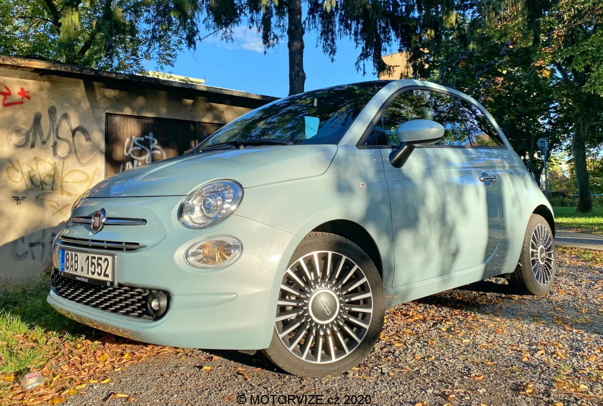 Foto en ángulo bajo de un Fiat 500 2015 desde el cuarto frontal, mostrando el lado del conductor. La imagen resalta la parrilla, el parachoques delantero, los faros, las luces antiniebla y las llantas de aleación detalladas. El coche está aparcado sobre una superficie cubierta de hojas otoñales, con una zona herbosa y una estructura grafiteada al fondo, iluminado por luz natural del día, sugiriendo iluminación de mañana o tarde.