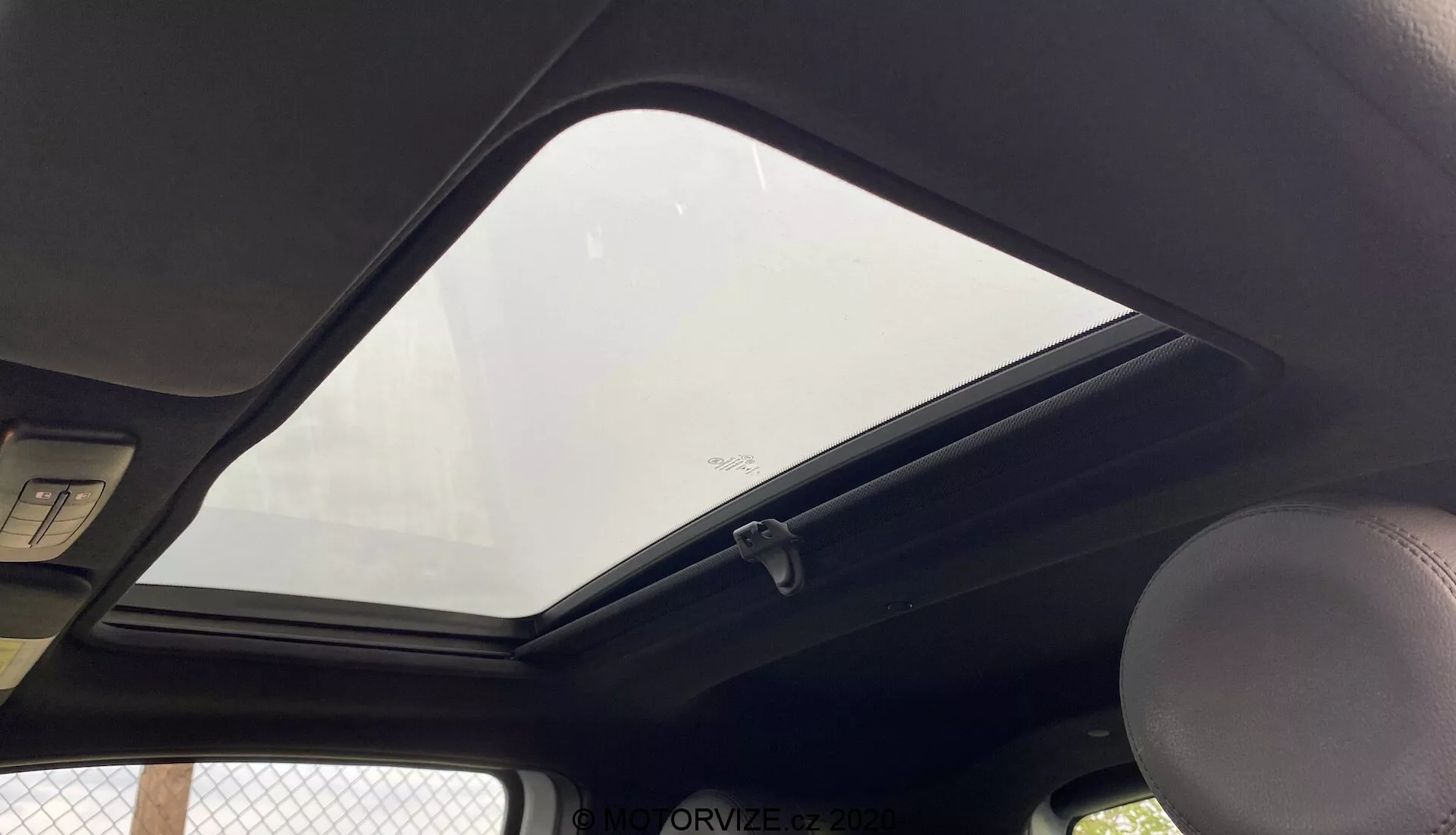 Vista interior de un Fiat 500 2015 enfocada en la sección superior hacia el techo desde la perspectiva de un pasajero o conductor. El prominente techo solar cerrado permite la entrada de luz natural en la cabina. El forro del techo de color oscuro contrasta con el exterior. Manija visible en el lado izquierdo, una luz de cúpula estándar encima y los bordes de dos viseras solares en la parte superior. Un gancho para ropa está cerca del borde del cristal del techo solar, que tiene una etiqueta de seguridad en la parte inferior. La imagen se toma desde un ángulo bajo, enmarcando de cerca las características del techo interior.