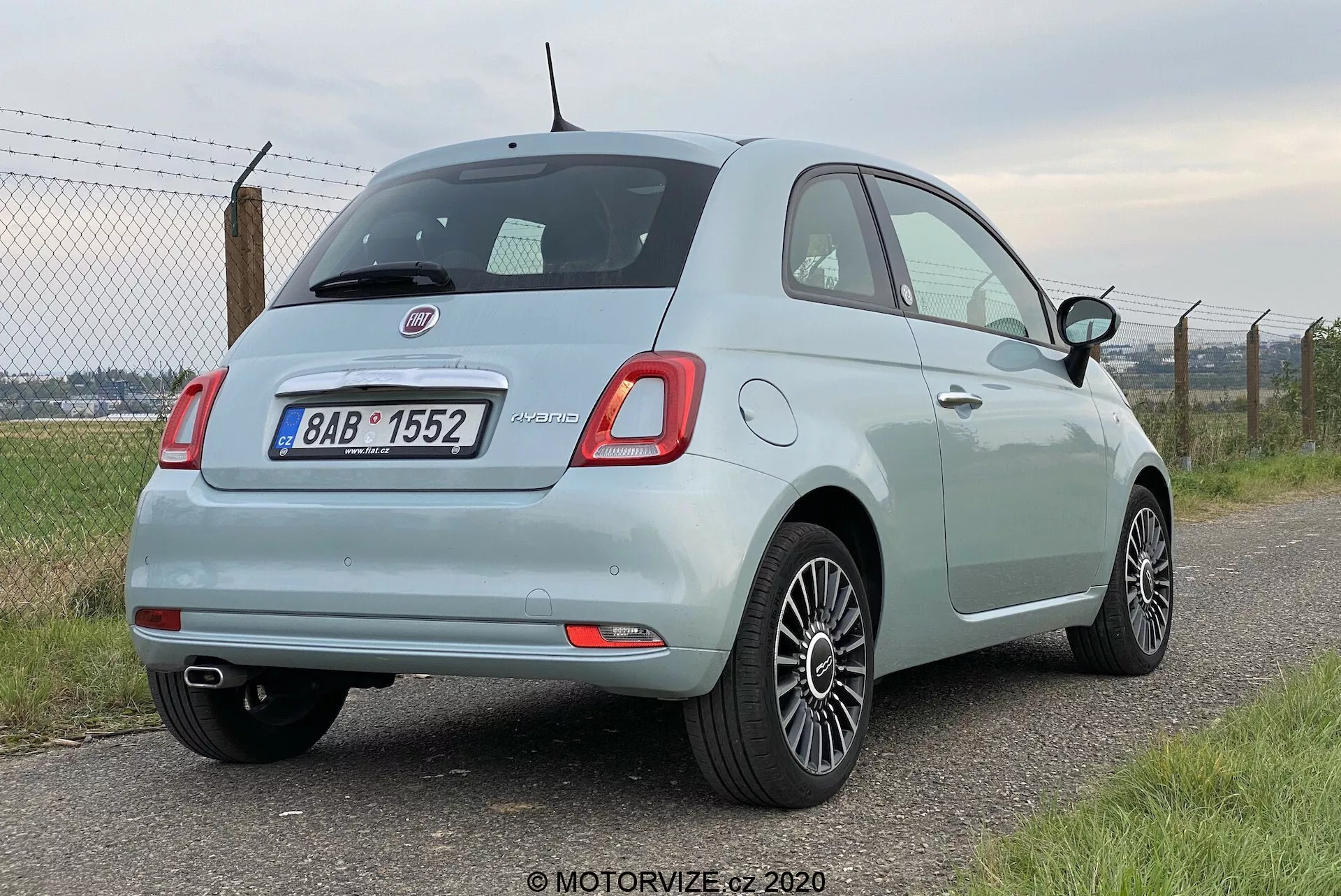 Vista de tres cuartos traseros de un coche compacto Fiat 500 de 2015 en color azul claro, que muestra sus luces traseras rectangulares redondeadas, limpiaparabrisas trasero, insignia cromada, sensores de aparcamiento en el parachoques, tubo de escape del lado izquierdo y llantas de aleación. El coche está aparcado con su lado izquierdo de cara a la cámara, frente a una valla y vegetación desenfocadas.