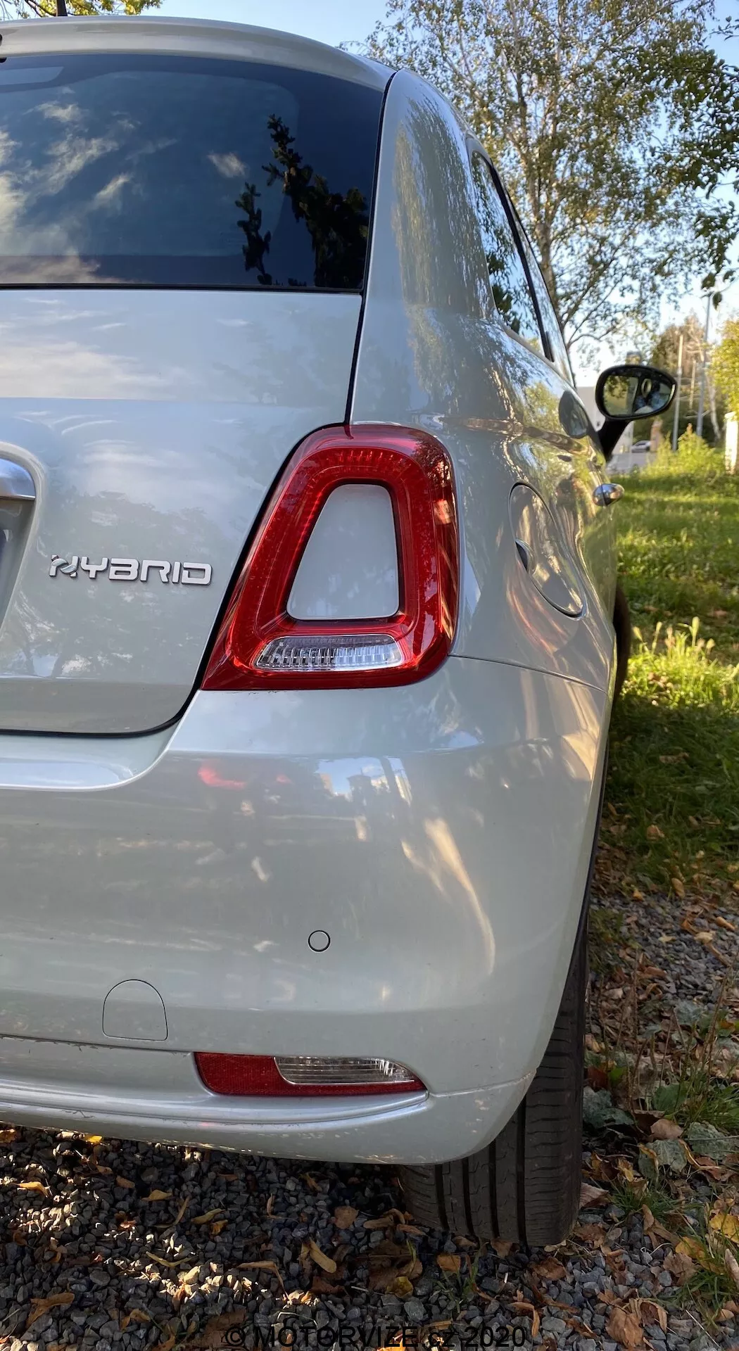 Vista trasera derecha de un Fiat 500 híbrido de 2015 en color plateado metálico, mostrando la luz trasera LED, la insignia híbrida, el parachoques trasero, una parte de la rueda de aleación, la ventana trasera tintada y la carrocería brillante; fotografiado en un entorno de parque suburbano con vegetación y árboles.