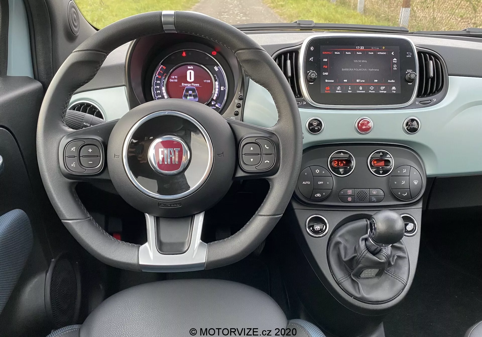 Vista interior de un Fiat 500 2015 desde la perspectiva del conductor mostrando el volante de tres radios con botones de control, cuadro de instrumentos digital parcialmente visible, pantalla táctil de infoentretenimiento apagada, botones y perillas de control climático, palanca de cambios manual, freno de mano y esquema de color de dos tonos enfatizando la funcionalidad y los elementos de diseño moderno.