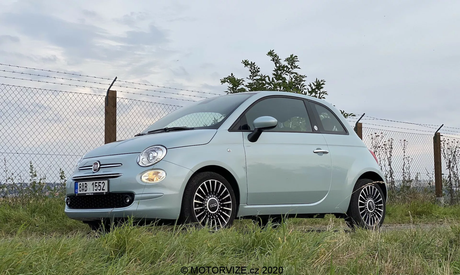 Foto en un ángulo bajo de un Fiat 500 de 2015, destacando su postura robusta. Vista de perfil lateral y parte delantera, con faros redondos, detalles cromados y emblema visible. Vehículo de dos puertas en color pastel estacionado en área de césped con fondo industrial o semiurbano. Ruedas de diseño multirradiado, espejos laterales del color de la carrocería y antena trasera visible. Parrilla frontal modesta con badge de Fiat en cromo.