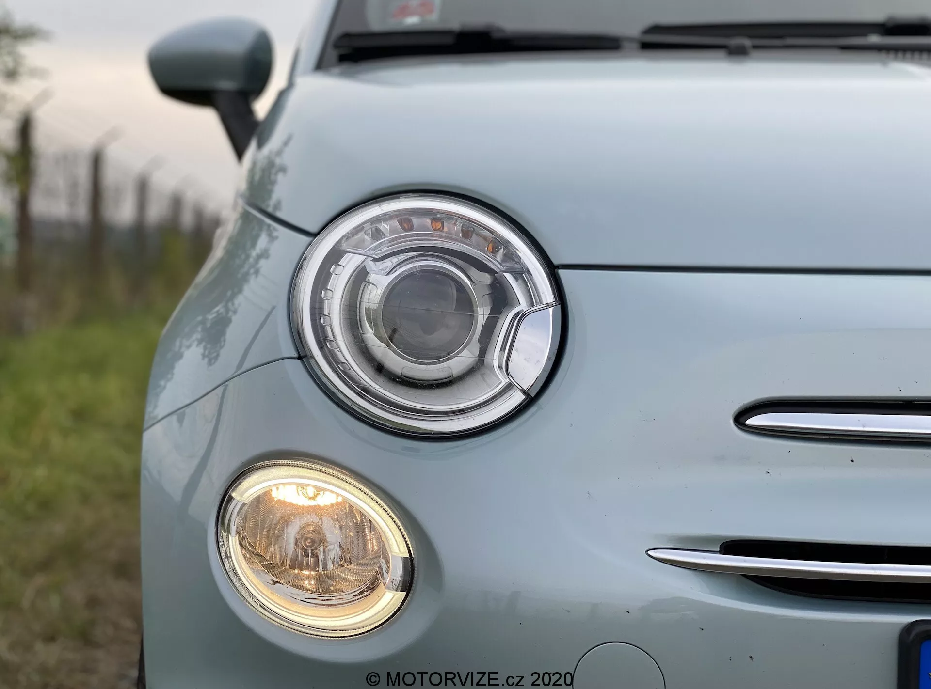 Imagen en primer plano de un Fiat 500 2015 azul claro capturada desde el cuarto delantero izquierdo. El enfoque está en el conjunto de faros distintivo con un diseño circular y lente transparente, así como en el faro antiniebla circular más pequeño debajo de él. A la izquierda se ve un segmento de la parrilla con listones horizontales cromados. La pintura azul pálido y brillante del coche refleja la luz natural, destacando su superficie bien mantenida. Las insignias y logotipos del vehículo han sido desdibujados.
