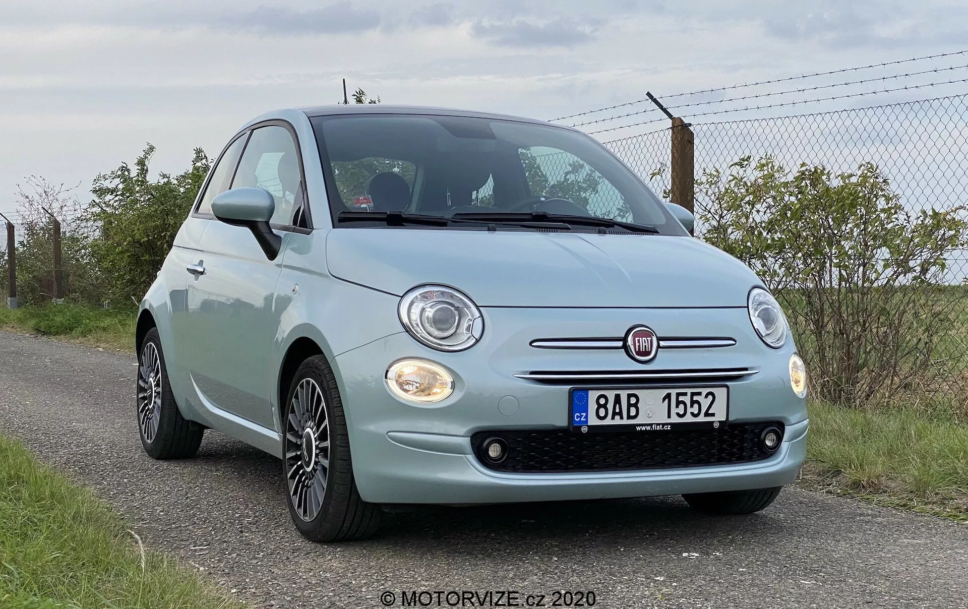 Vista frontal en ángulo bajo de un Fiat 500 de 2015, destacando la fascia delantera, los faros redondos, los detalles cromados de la parrilla, las luces antiniebla y el emblema prominente de Fiat. El capó del coche presenta curvas características y el parachoques delantero incluye sensores de estacionamiento visibles. También son visibles las llantas de aleación. El vehículo está estacionado al borde de la carretera con una zona de césped y una valla en el fondo, indicando un entorno rural o semi-rural.