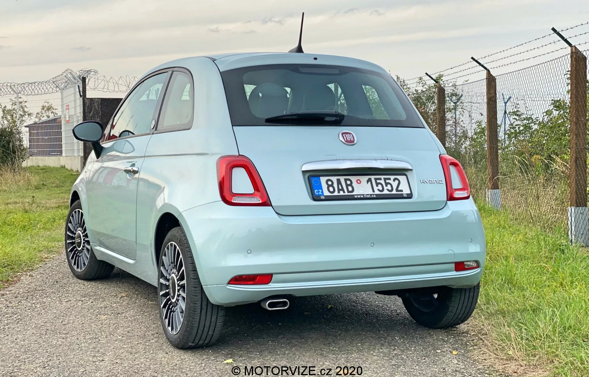 La imagen muestra una vista de tres cuartos trasera de un coche hatchback Fiat 500 del 2015 de color azul claro. El enfoque está en su parte trasera y parte de su perfil lateral, tomado desde el lado trasero derecho del vehículo. Destaca la rueda trasera derecha, la luz trasera, la vista parcial del espejo lateral derecho y la puerta del pasajero. El coche está ligeramente colocado a la izquierda, con el ángulo de la cámara ligeramente más bajo que la línea del techo. Detalles notables incluyen una punta de escape única, ruedas de aleación, luces traseras de bombilla estándar, insignias de marca y modelo, un pequeño alerón trasero, una antena estilo mástil y una tapa de combustible visible. El entorno parece rural con cielos abiertos, vegetación y una cerca.