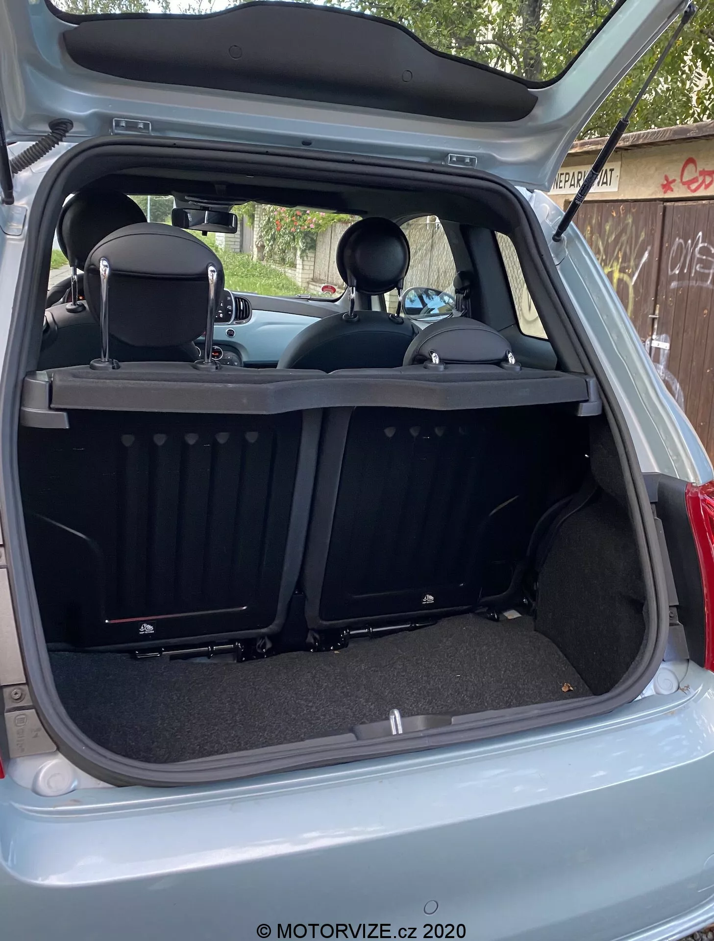 La imagen muestra el interior trasero del Fiat 500 2015 desde un ángulo trasero externo con la puerta del maletero completamente abierta. El área de carga se ve desde un ángulo ligeramente descentrado y hacia abajo desde el lado derecho, revelando la profundidad del maletero y varias características interiores traseras. El diseño moderno y limpio incluye superficies alfombradas, una tapa de carga retráctil acanalada, asientos traseros abatibles divididos con reposacabezas y una placa de desgaste. Las características tecnológicas incluyen una luz interior, una toma de corriente de 12V, molduras plásticas, recortes circulares y paneles laterales.