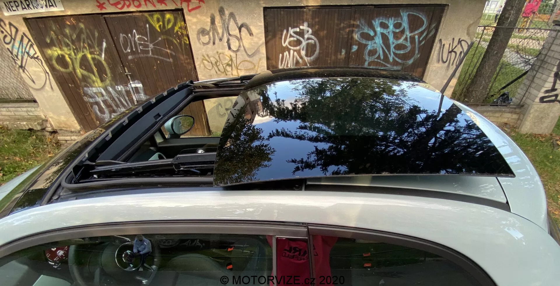 Vista trasera de primer plano de un Fiat 500 2015 centrada en la parte superior trasera, incluyendo el gran parabrisas trasero tintado envolvente, un alerón deportivo, un techo solar/panorámico, y una tapa de maletero abierta, reflejándose el verdor exterior y el cielo diurno.