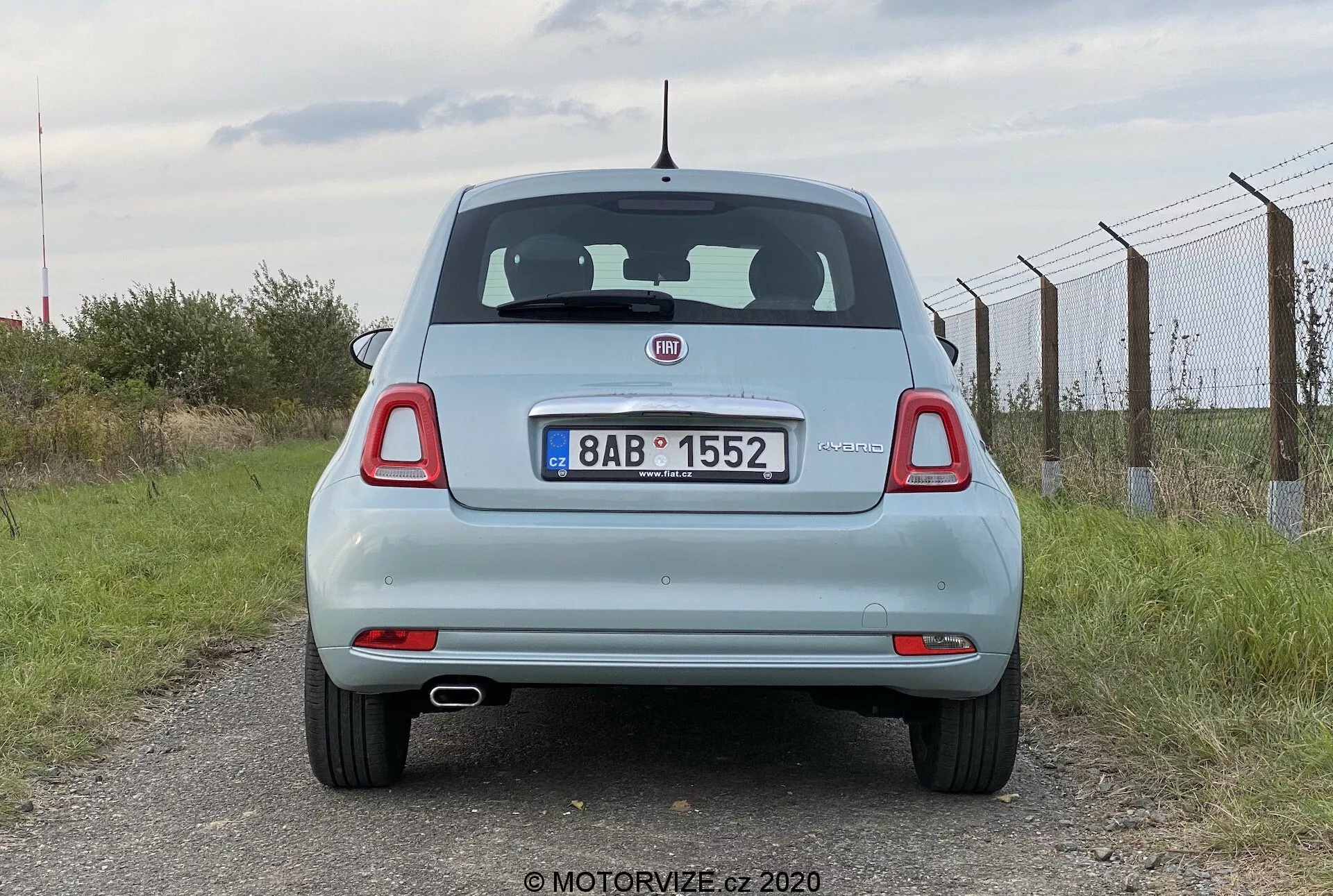 Vista trasera de un automóvil Fiat 500 2015 de color azul claro. La perspectiva a nivel de los ojos captura el vehículo estacionado, mostrando toda su parte trasera, incluyendo la tapa del maletero, parabrisas trasero, luces traseras, parachoques trasero y placa de matrícula. Las características incluyen el emblema del fabricante prominente, limpiaparabrisas trasero, luz de freno montada alta y luces traseras redondas con segmentos adicionales. La placa de matrícula está montada en el centro de la tapa del maletero, debajo del emblema y por encima del parachoques trasero plano con reflectores integrados. Una antena corta está posicionada en el centro del techo hacia la parte trasera. Cielo nublado y entorno rural, con una valla a la derecha y césped debajo.