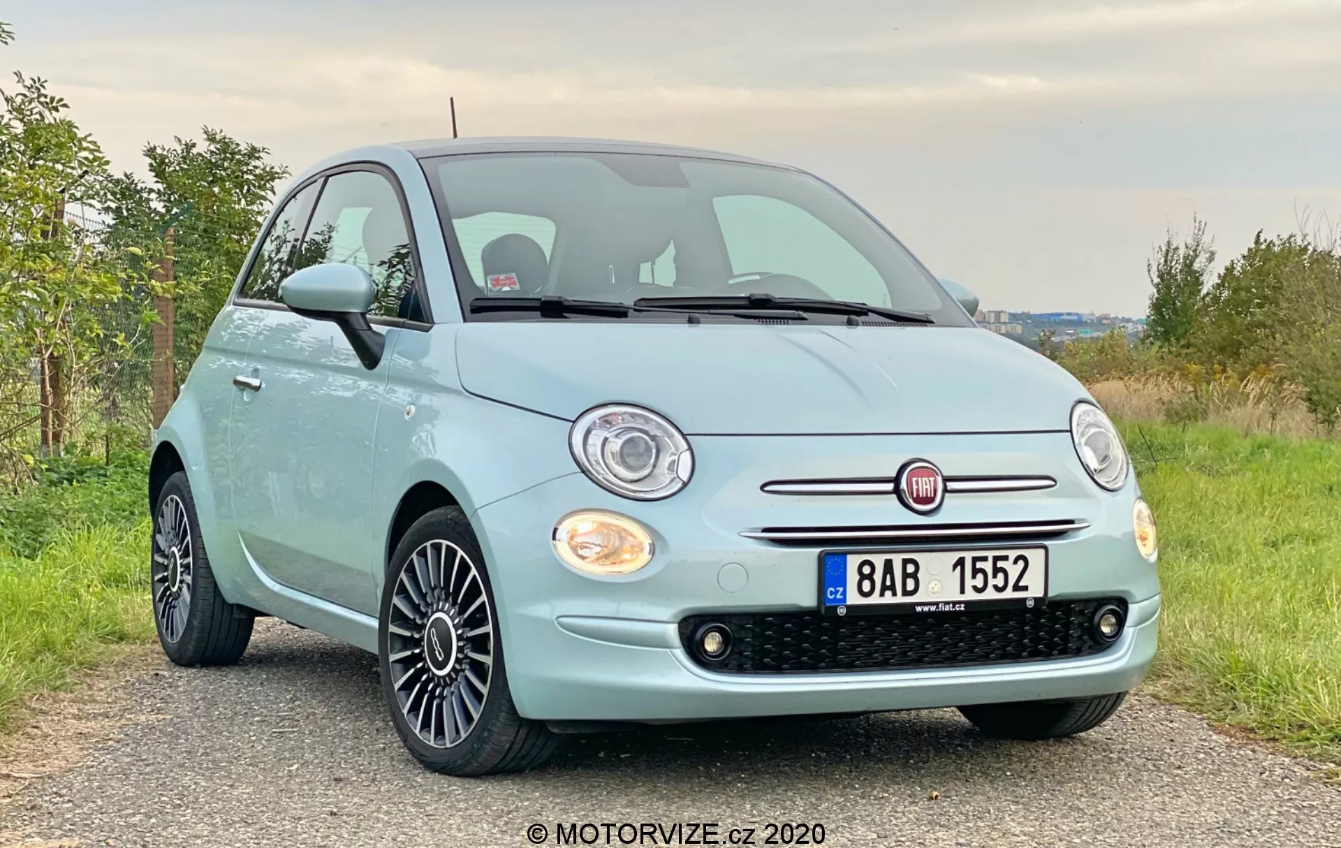 Vista frontal de tres cuartos de un Fiat 500 azul pálido de 2015 con rejilla acentuada en cromo y faros halógenos redondos en un ángulo ligeramente bajo. La imagen resalta la fascia frontal, la rejilla, los faros, el capó y las llantas de aleación con diseño de múltiples radios negros. El coche está estacionado en un camino de grava o tierra en una zona rural con vegetación verde en el fondo.