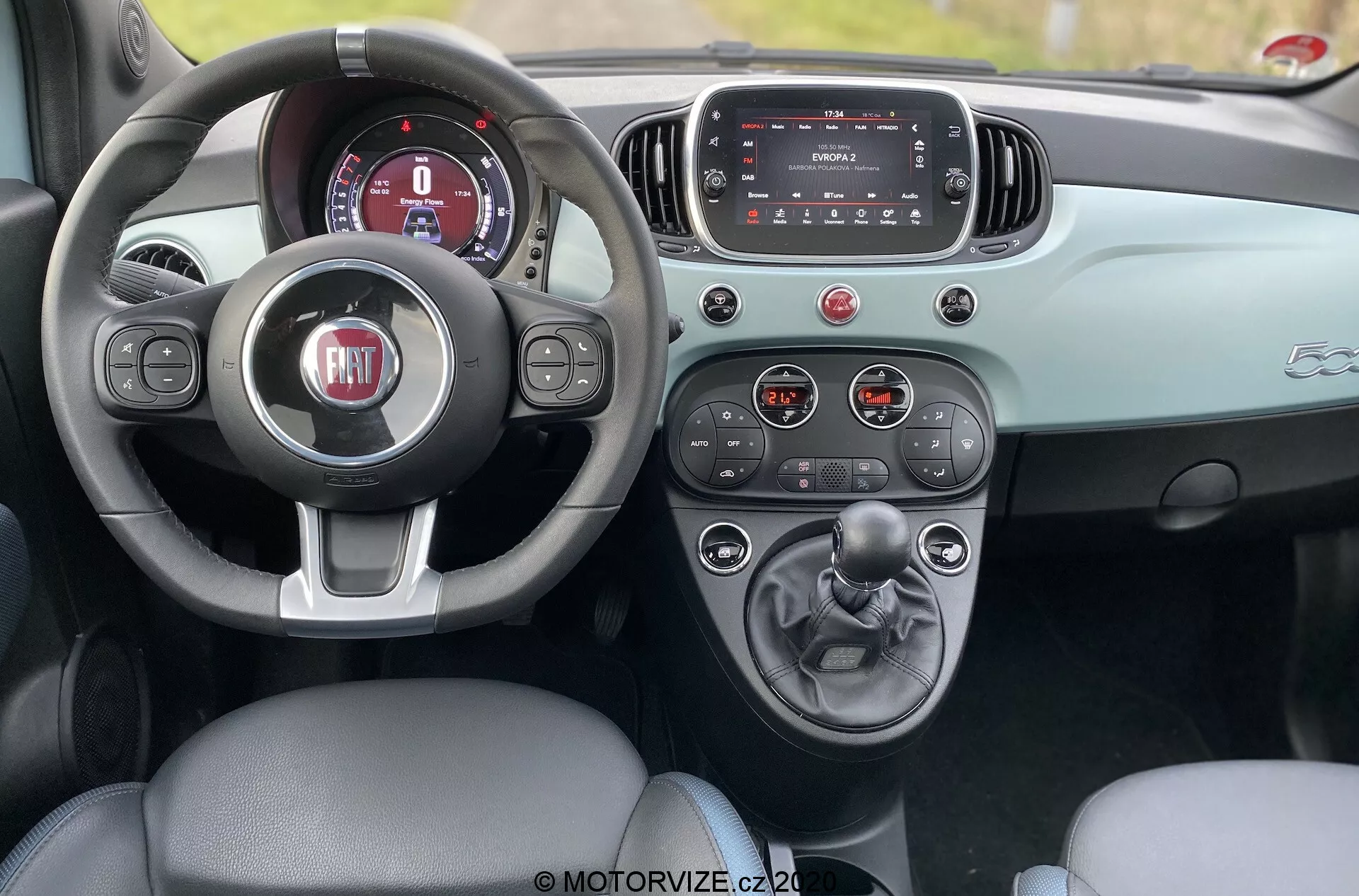 Toma interior de un Fiat 500 de 2015 desde el área de asientos traseros, mostrando la cabina delantera con un asiento de cuero para el conductor, volante de tres radios con el logotipo de FIAT, panel de instrumentos digital, pantalla táctil de infoentretenimiento y consola central con cambio manual y soportes para vasos. El tablero y la puerta tienen un esquema de color en dos tonos que realza el diseño estético. La foto fue tomada durante el día, resaltando el diseño interior refinado y la funcionalidad del coche.