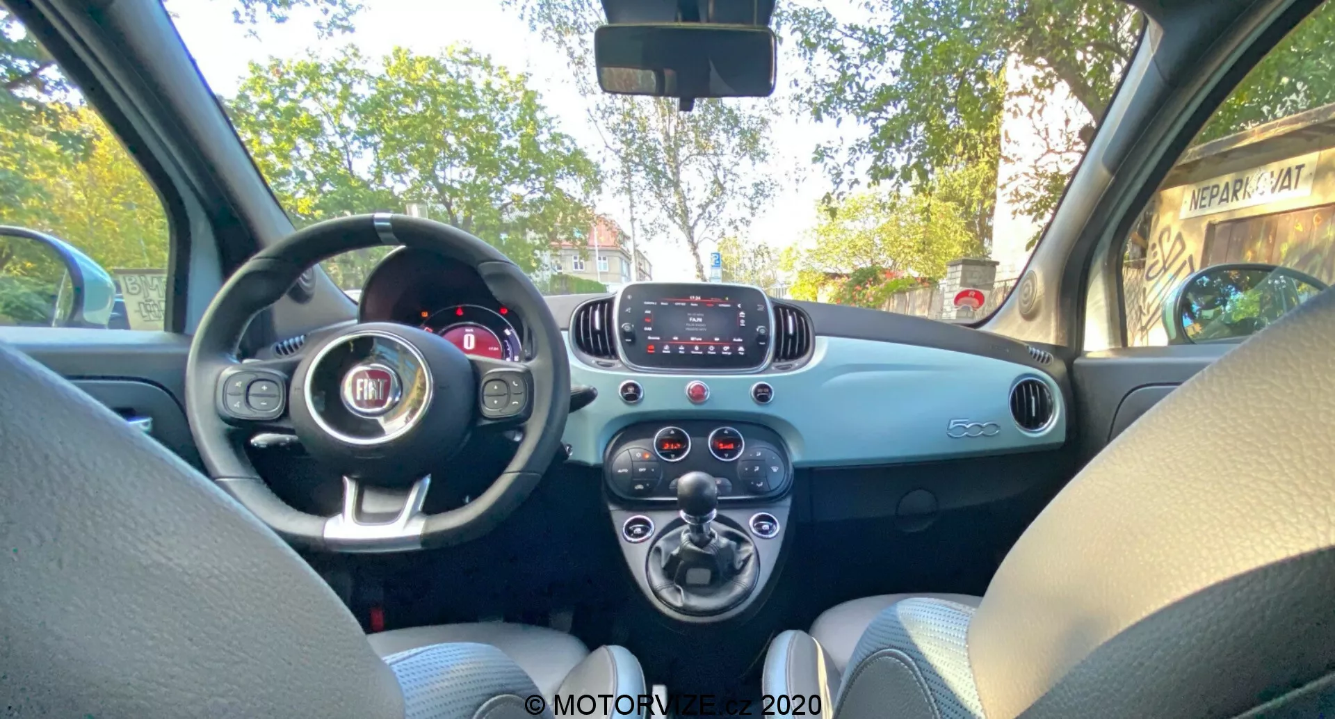 Vista interior del Fiat 500 2015 desde el asiento trasero, mostrando el tablero frontal, volante con el emblema de Fiat, cuadro de instrumentos parcialmente visible, sistema de infoentretenimiento básico con botones, diales de control del clima, tablero de dos tonos, asientos de tela gris claro, puerta del lado del pasajero con manija y rejilla del altavoz, diseño simple y funcional sin características tecnológicas de alta gama.