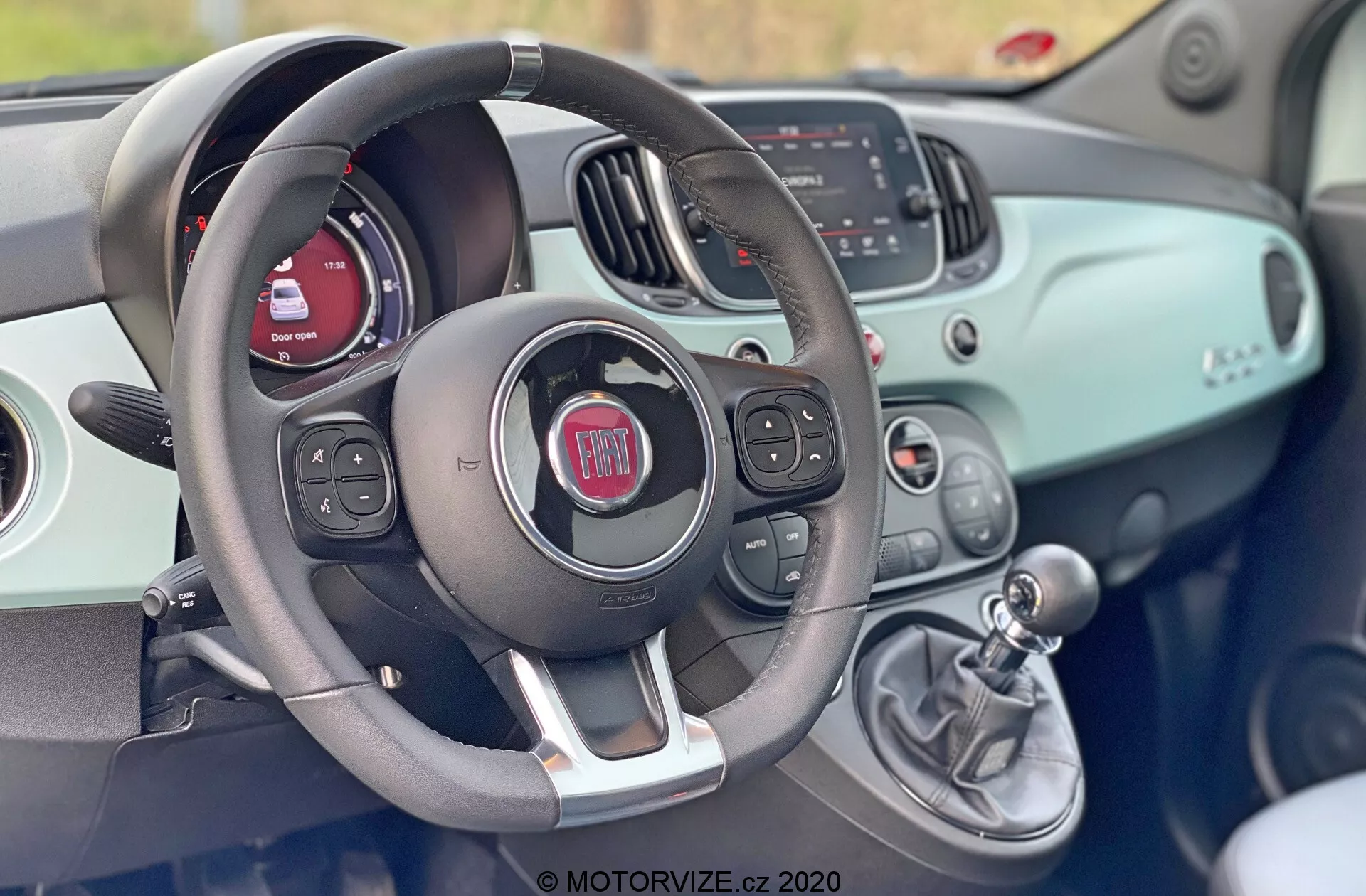 Vista interior de un Fiat 500 de 2015 desde el asiento del conductor que muestra el volante con controles integrados para audio, comandos de voz y posiblemente control de crucero. Detrás del volante, el grupo de instrumentos presenta una pantalla digital y medidores tradicionales. El tablero tiene una superficie del mismo color que la carrocería y cuenta con una pantalla de infoentretenimiento que muestra información de radio y medios, así como controles manuales de HVAC. A la derecha del volante, una palanca de cambio manual indica una transmisión manual. El diseño general es moderno con una estética deportiva y bicolor.