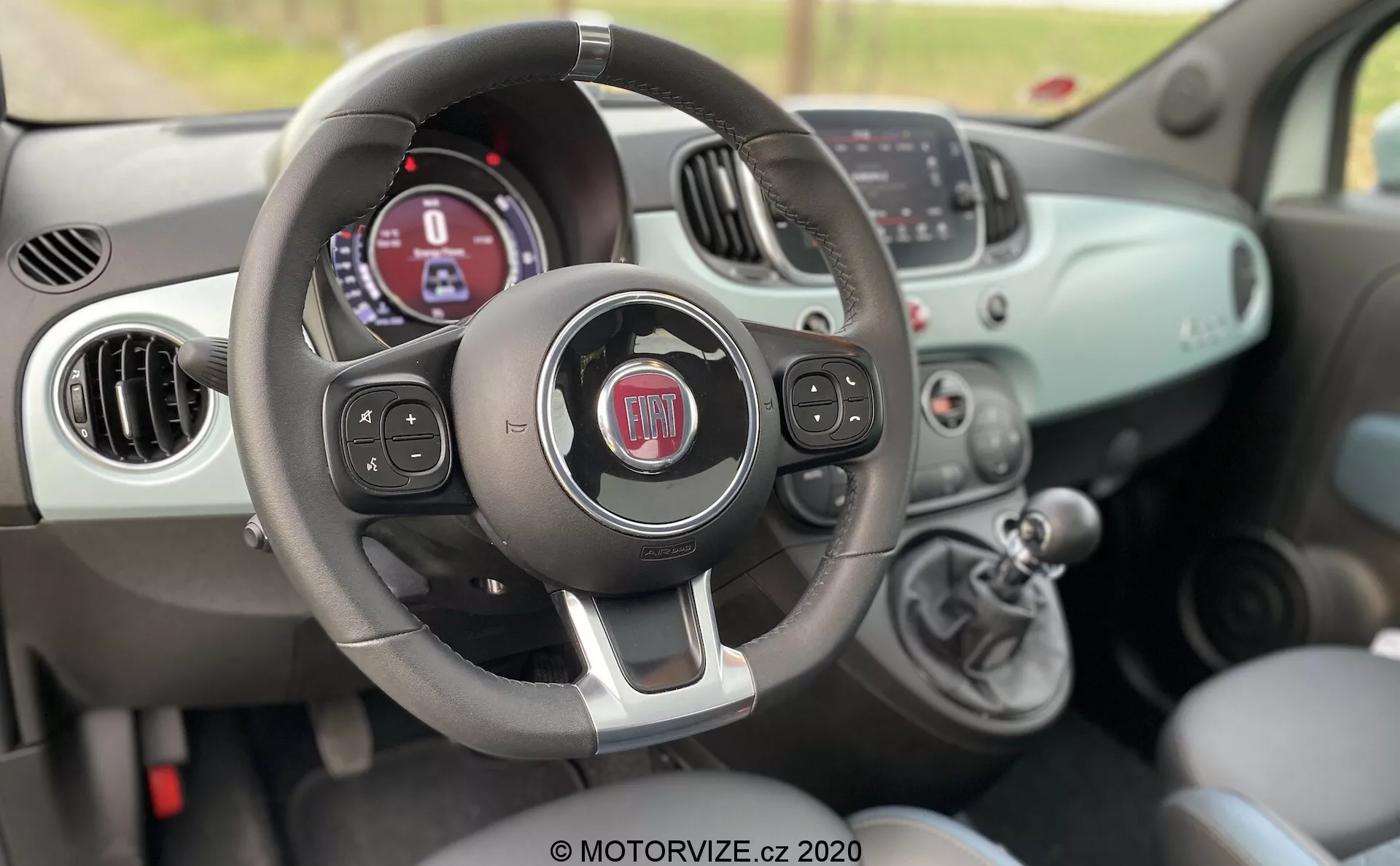 Vista interior de un Fiat 500 de 2015 desde el lado del pasajero, mostrando el volante con botones multifunción, el panel de instrumentos con pantalla digital, los controles de clima manual y la palanca de cambios.