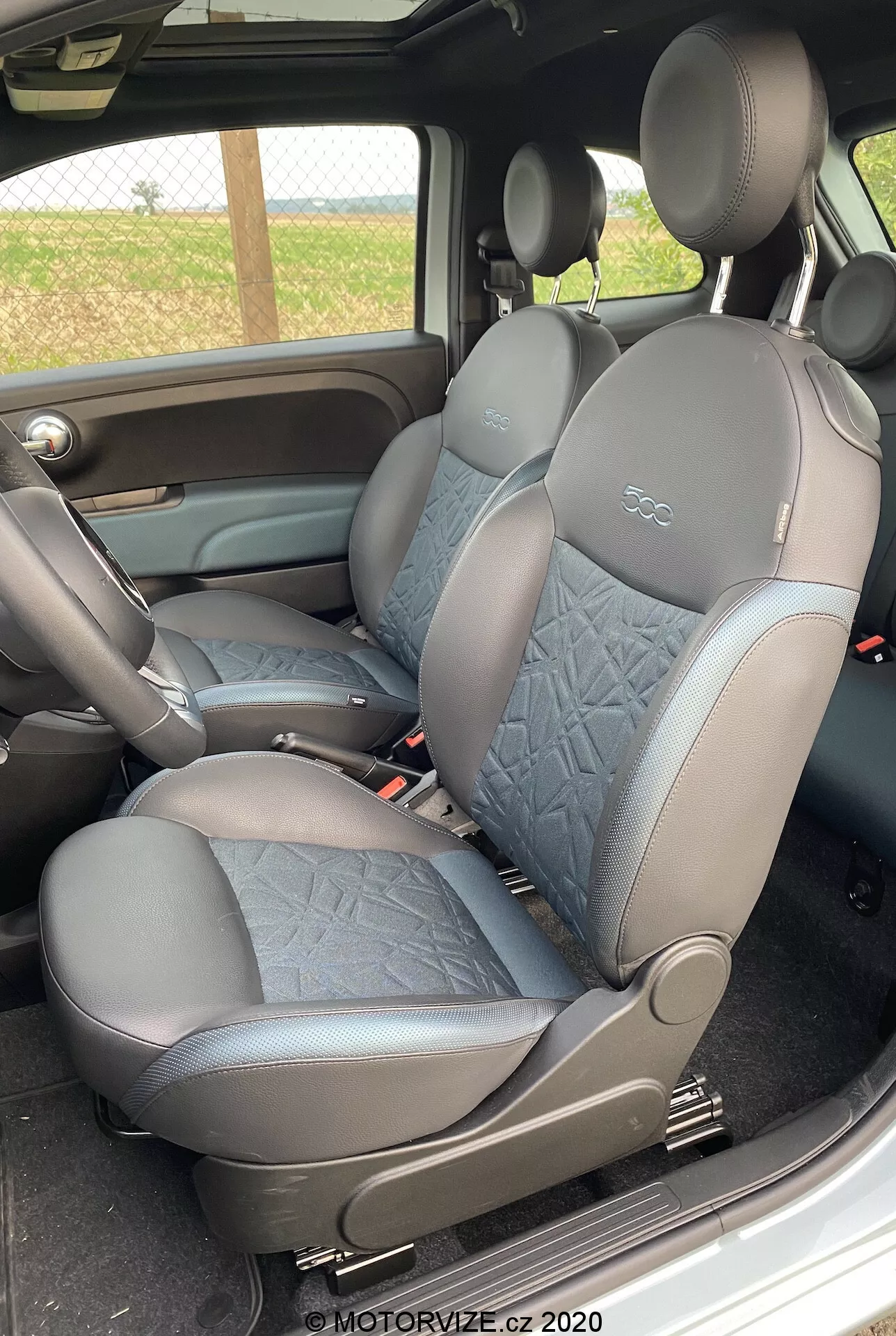 La imagen muestra el interior de un Fiat 500 2015 en la zona de pasajeros, con vista lateral de los asientos delanteros. Tapicería de cuero y tela acolchada. Reposacabezas integrados. Tono oscuro por su practicidad. Palanca de ajuste manual de asiento visible. Apoyabrazos de la consola central parcialmente visible. Coincidencia de color y material con el panel de la puerta. Marca de agua de 2020.