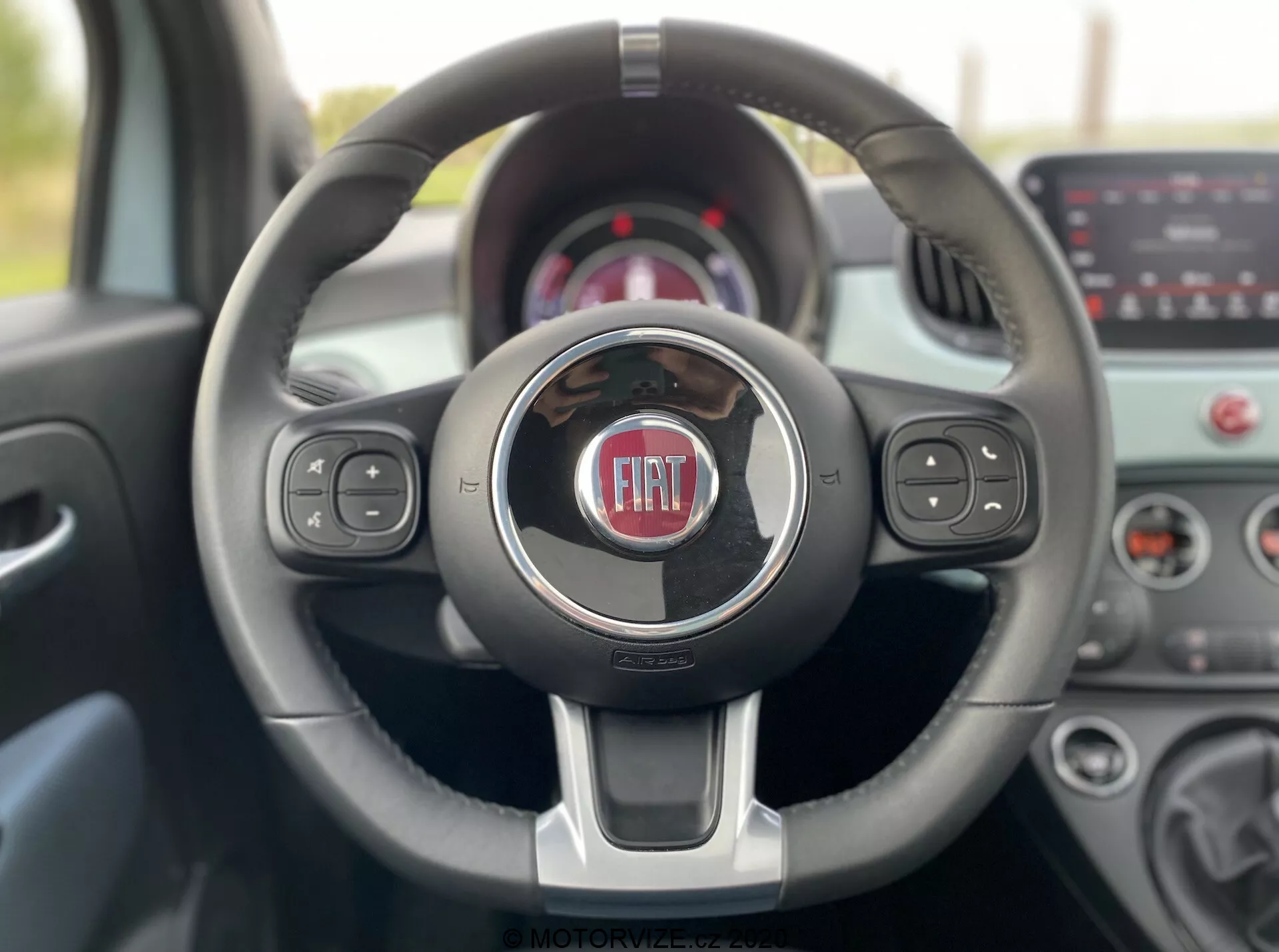 Vista del interior de un Fiat 500 2015 desde el asiento del conductor, mostrando un volante moderno de tres radios con acentos de cromo y el logotipo de Fiat en el centro. El volante incluye controles integrados para medios, funciones de voz y control de crucero. El fondo borroso presenta un cuadro de instrumentos y un sistema de infoentretenimiento con pantalla táctil, destacando la tecnología y diseño contemporáneos del vehículo.