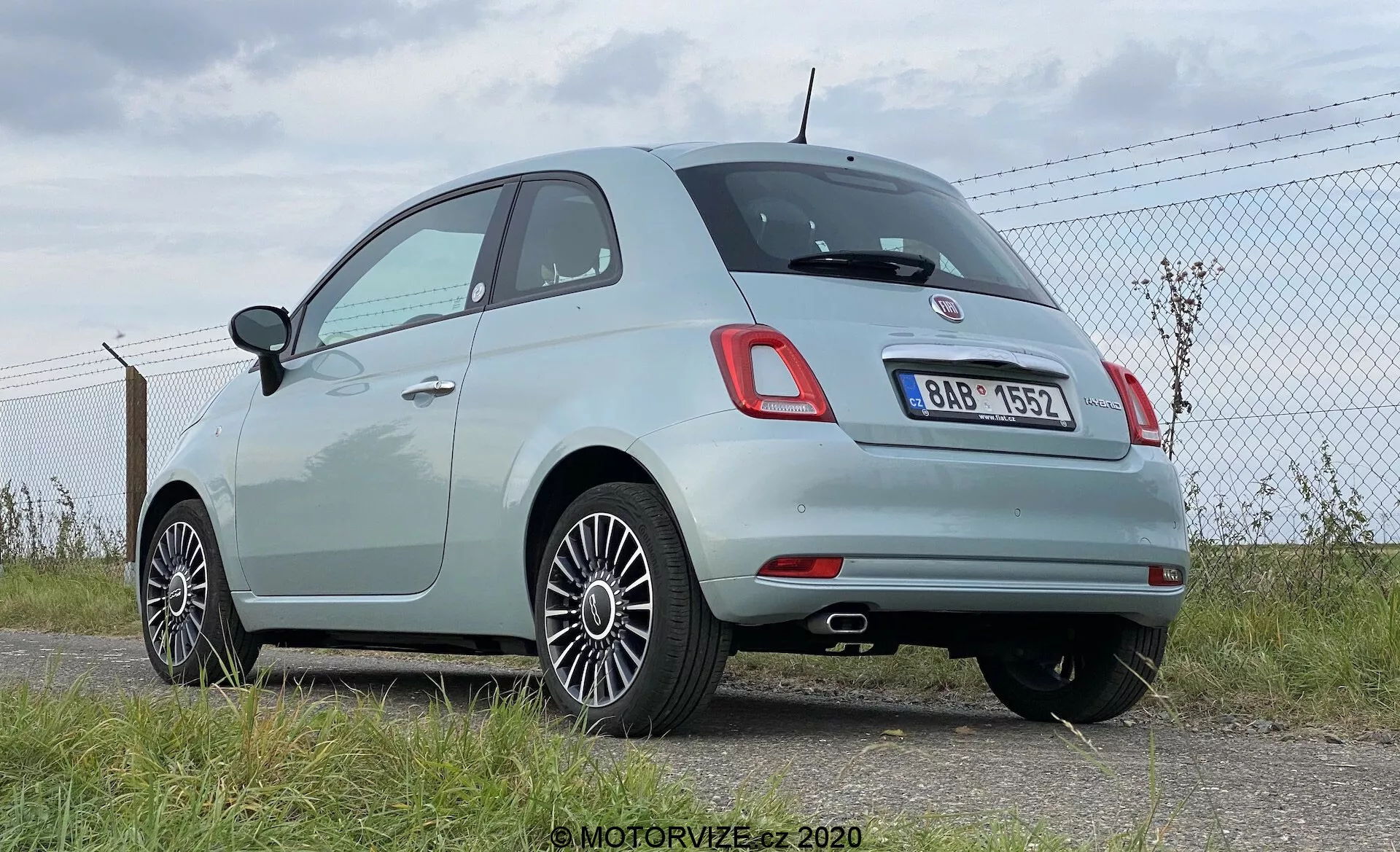 Vista trasera tres cuartos de un moderno Fiat 500 hatchback de color claro, que muestra luces traseras redondeadas distintivas y una insignia central cromada encima del área de la placa de matrícula trasera. La imagen resalta la línea del techo redondeada del vehículo, el borde cromado alrededor de las ventanas laterales y las llantas de aleación multirradio. La pintura pastel clara del automóvil indica que está relativamente nuevo o bien mantenido. Está estacionado cerca de una cerca de alambre en un área pavimentada, con el cielo y el horizonte visibles en el fondo, lo que sugiere un aeródromo o un área abierta. Las posibles características tecnológicas incluyen sensores de estacionamiento traseros integrados en la sección negra del parachoques trasero.