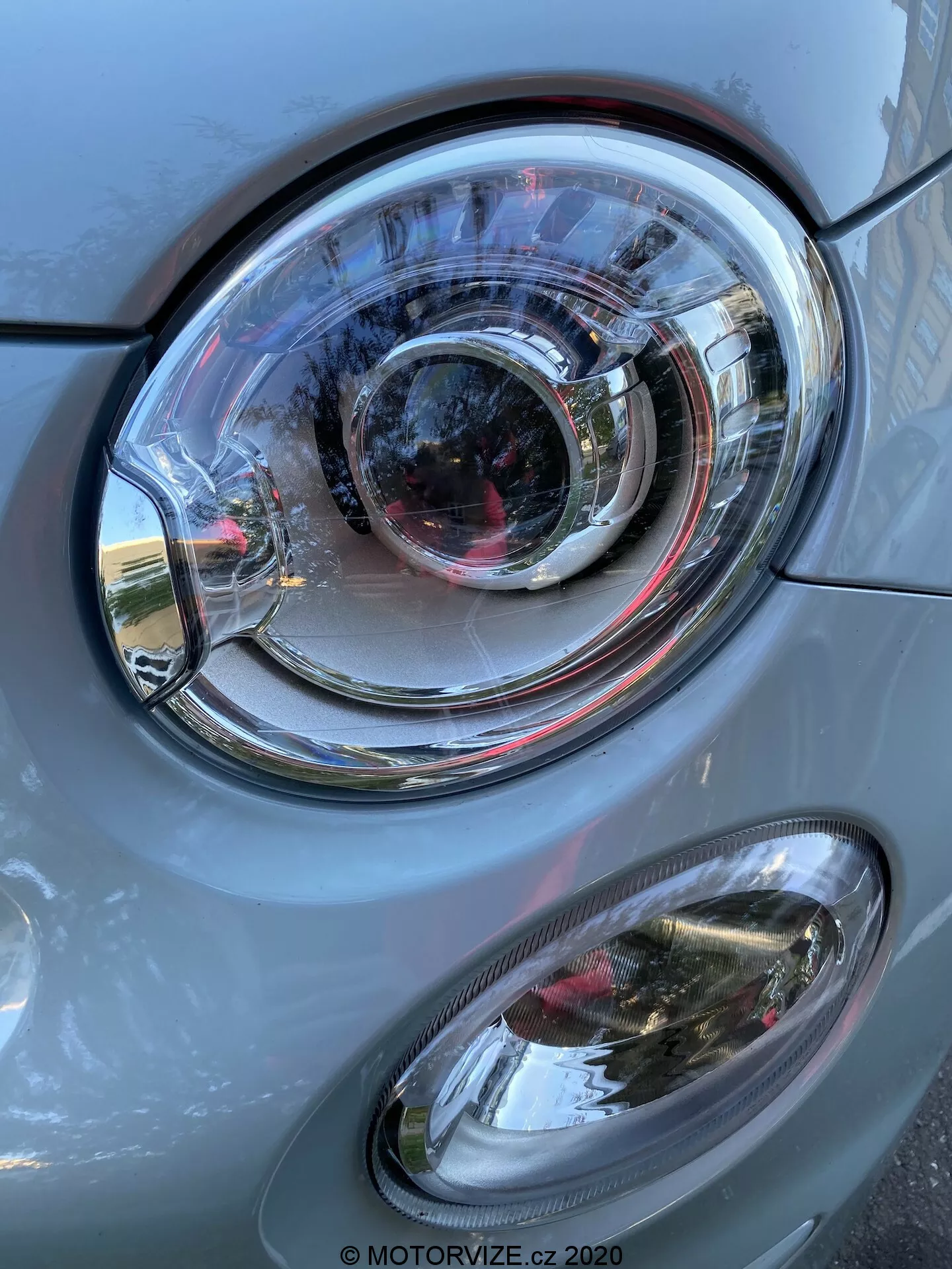 Vista en primer plano del lado izquierdo delantero de un coche Fiat 500 2015, enfocándose en el ensamblaje del faro. La imagen, en orientación vertical, presenta un prominente faro principal de lente clara de policarbonato con un diseño complejo de anillos concéntricos, indicando un estilo de proyector y posiblemente tecnología de iluminación adaptativa. Debajo del faro principal hay una lámpara antiniebla secundaria ovalada con una cubierta clara. Los paneles de la carrocería del coche, de color gris con un brillo metálico, tienen un acabado liso, reflejando la luz del día y elementos del entorno como edificios y cielo. El ensamblaje del faro y la lámpara antiniebla están en buen estado, indicando un vehículo bien mantenido o nuevo.