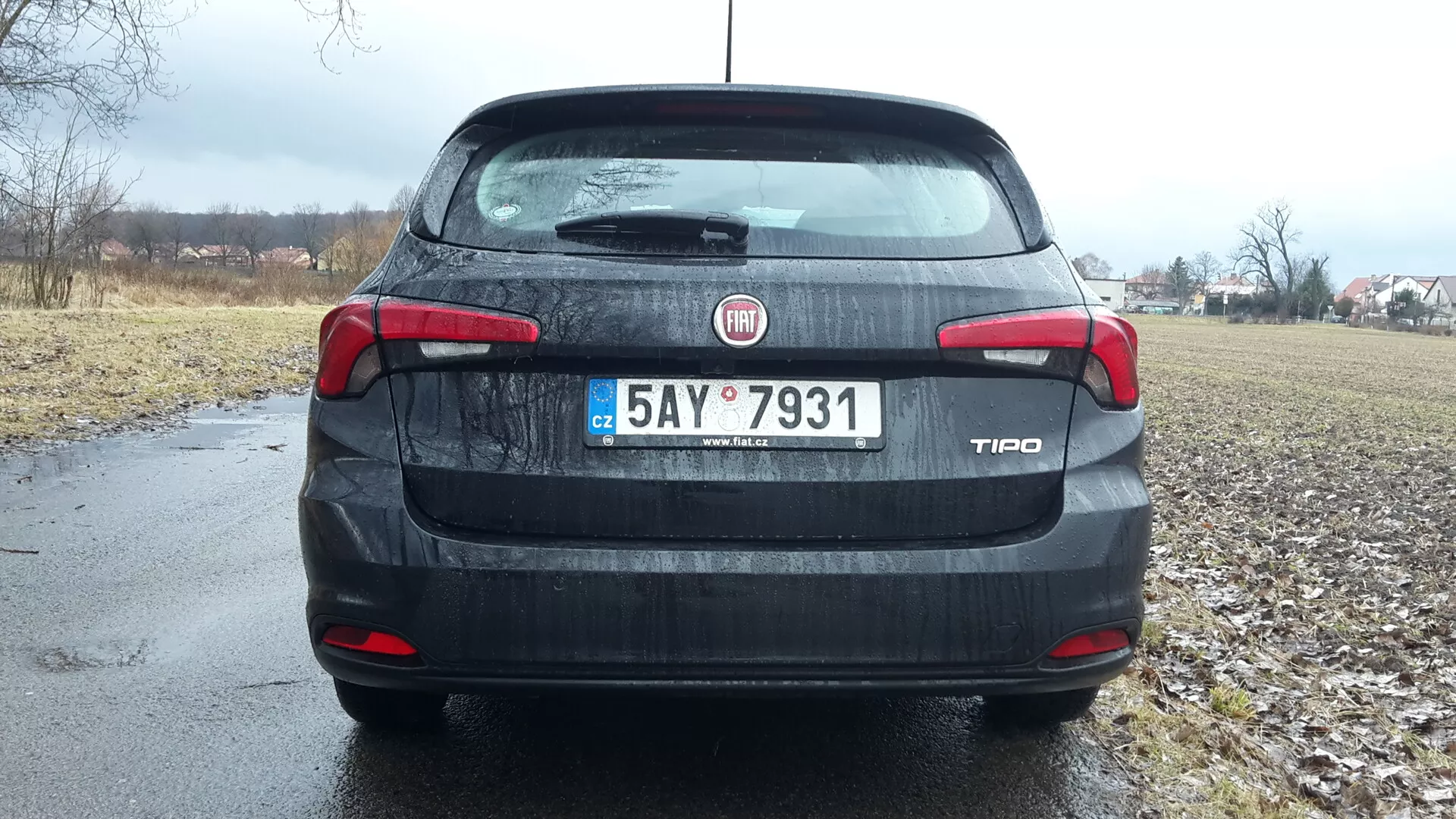 Vista trasera de un sedan Fiat Tipo 2016 bajo cielos nublados, con identificación de marca y modelo en el maletero, luces traseras y tercera luz de freno detalladas, placa de matrícula y un atisbo del entorno rural en el fondo