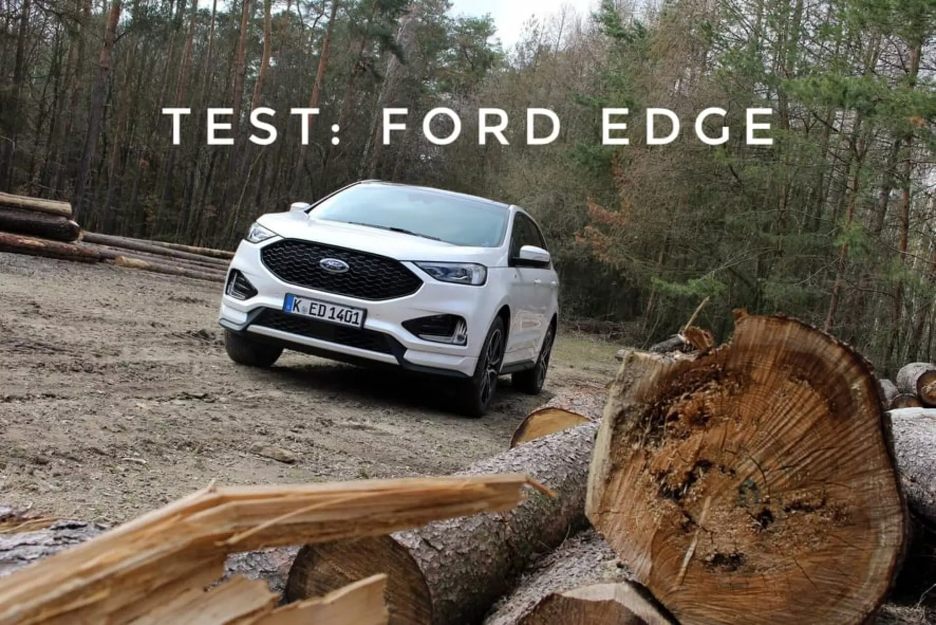 Ford Edge 2018 en blanco, demostrando su capacidad todoterreno con una vista frontal y lateral de tres cuartos, resaltando las luces diurnas LED, ruedas de aleación de múltiples radios y pintura bitono, ambientado contra un telón de fondo natural con troncos de madera cortada en primer plano
