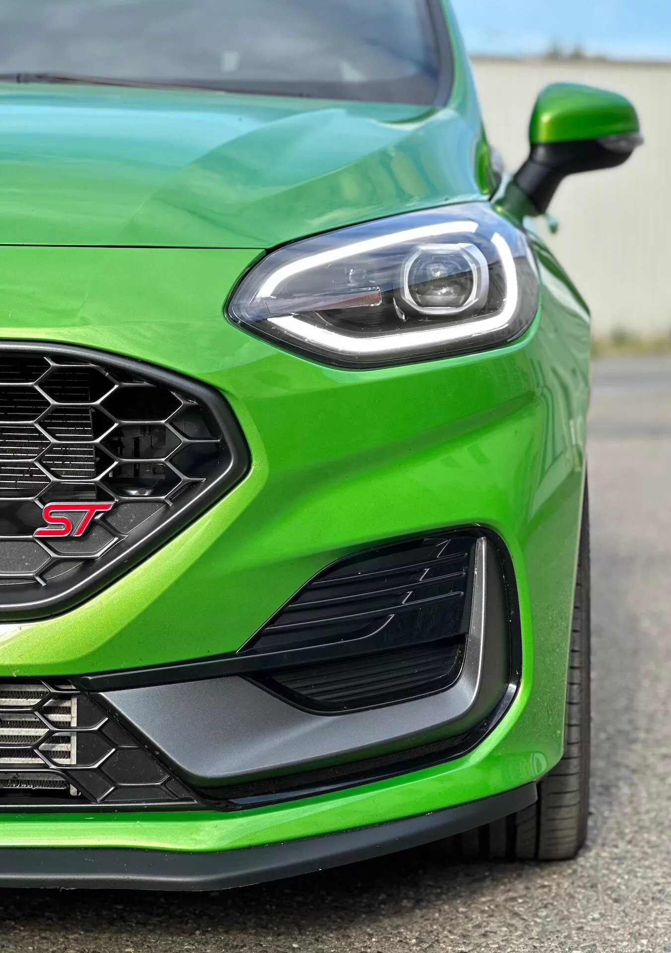 Vista detallada del cuarto frontal izquierdo de un Ford Fiesta ST 2022 en verde vibrante, centrada en el acabado de alto rendimiento, con rejilla de patrón de panal para flujo de aire óptimo, diseño avanzado de faros LED, ventilación lateral aerodinámica y parachoques delantero para mejorar la canalización del aire.