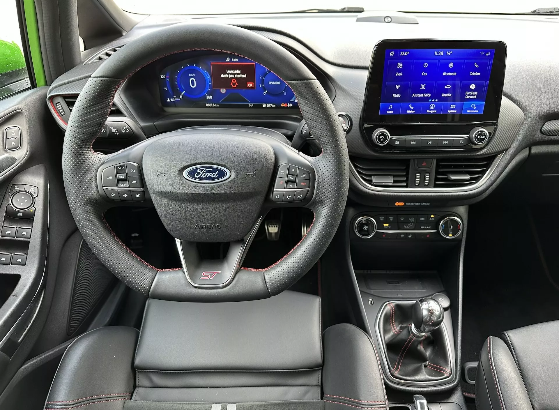 Vista interior del Ford Fiesta 2022 desde el asiento trasero, destacando el volante con el logo de Ford y la insignia ST, panel de instrumentos digital, sistema de infoentretenimiento táctil, palanca de cambios manual y asiento del conductor deportivo con costuras en contraste.