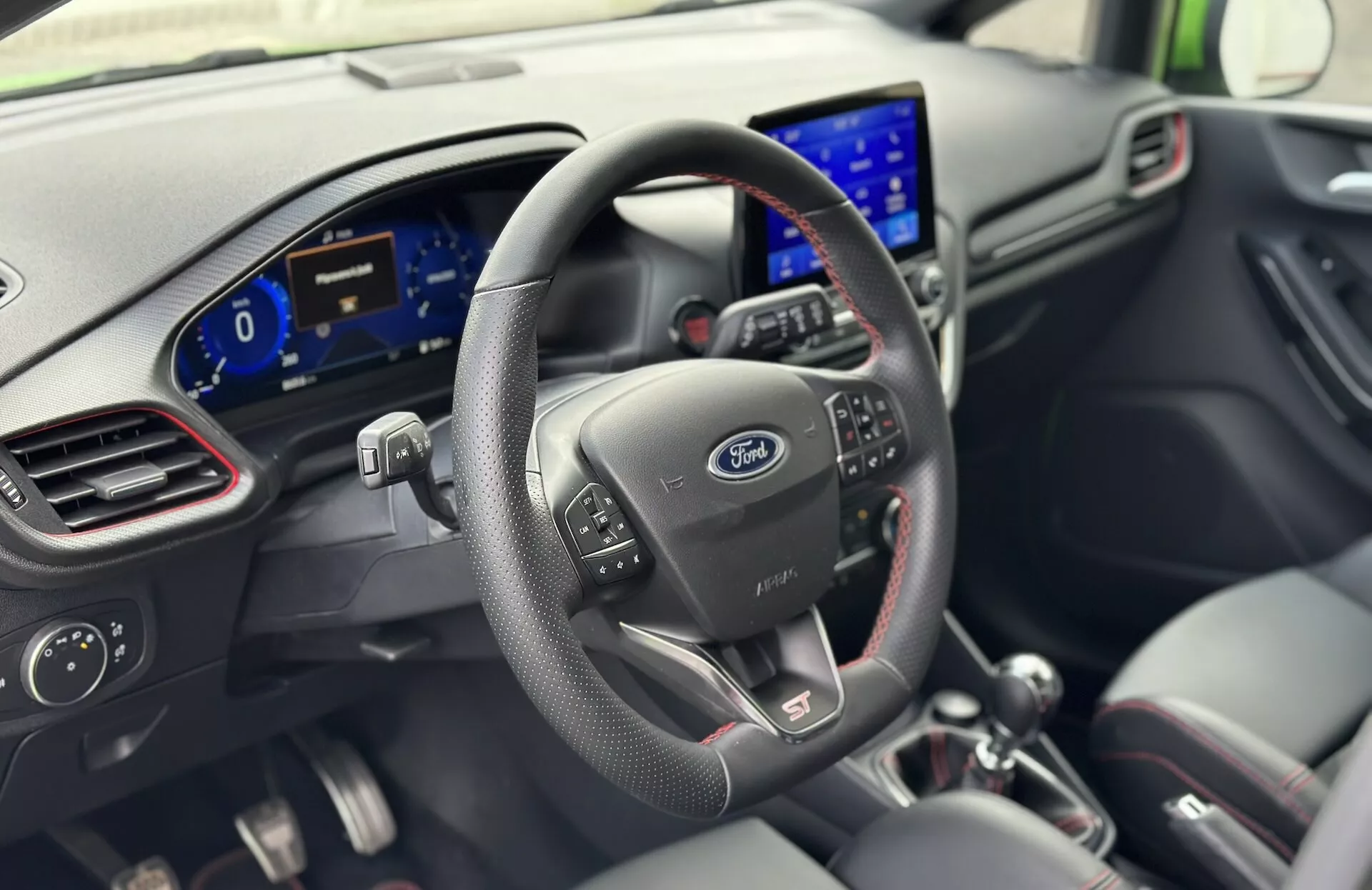 Interior del Ford Fiesta 2022 desde la perspectiva del conductor con volante de cuero con botones de control, logo de Ford, cuadro de instrumentos digital y sistema de infoentretenimiento moderno en el tablero