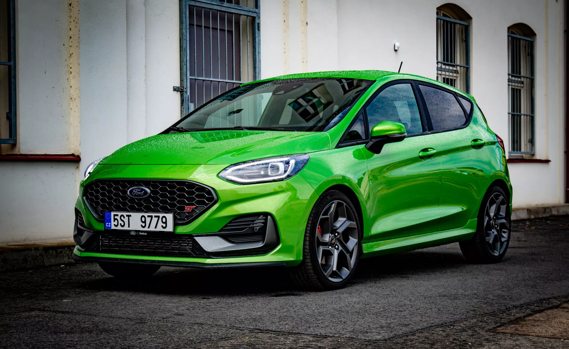 Ford Fiesta 2022 en verde brillante, mostrando su diseño deportivo desde un ángulo bajo de tres cuartos delantero resaltando el parachoques delantero, la parrilla, los faros, el capó, el guardabarros, las ruedas y el reflejo del parabrisas en una calle urbana después de la lluvia