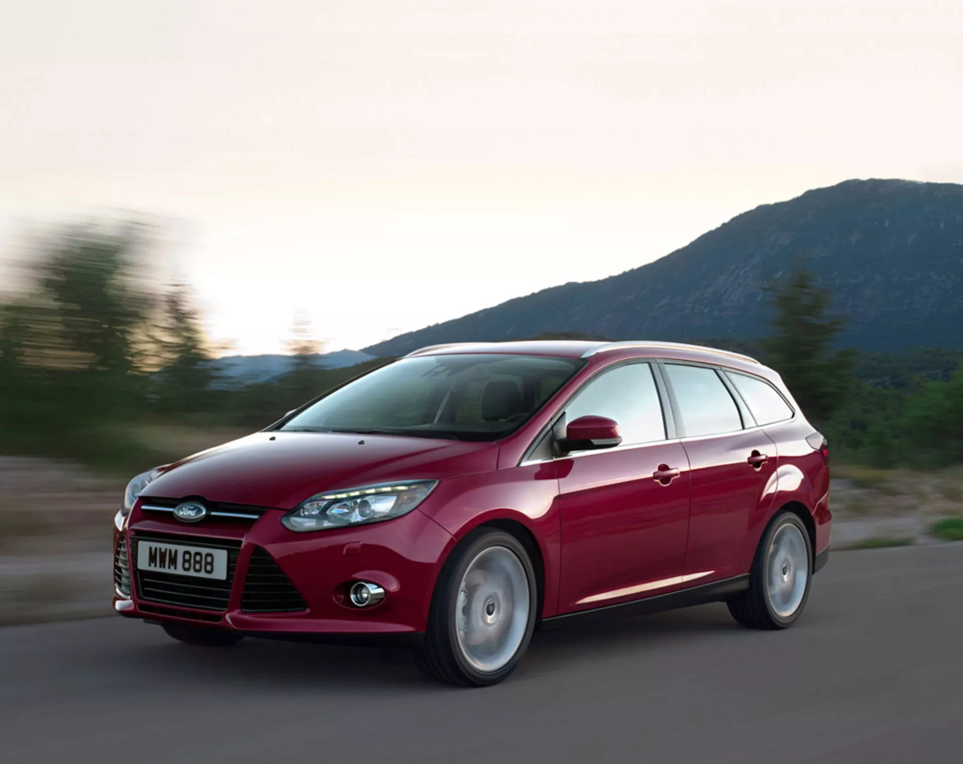 Ford Focus station wagon rojo 2011 en movimiento sobre una carretera de asfalto, mostrando su diseño moderno, parrilla frontal prominente con el logo de Ford, faros inclinados hacia atrás, líneas esculpidas en el capó y ruedas de aleación, bajo condiciones de iluminación vespertina