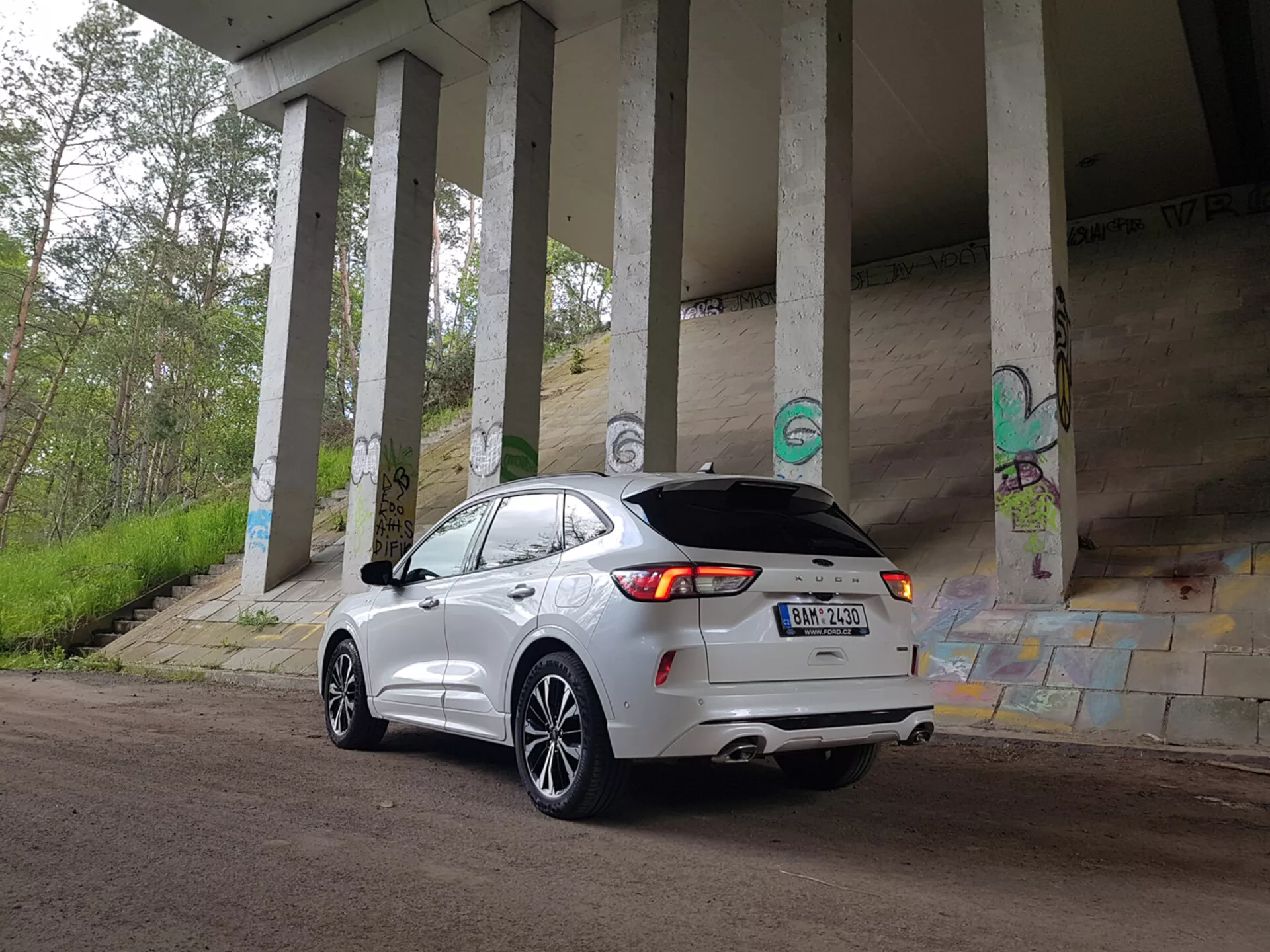 SUV crossover Ford Kuga 2019 estacionado debajo de un paso elevado, mostrando su diseño moderno y características tecnológicas como luces traseras LED, doble escape y cámara trasera, entre grafitis urbanos y pilares de concreto
