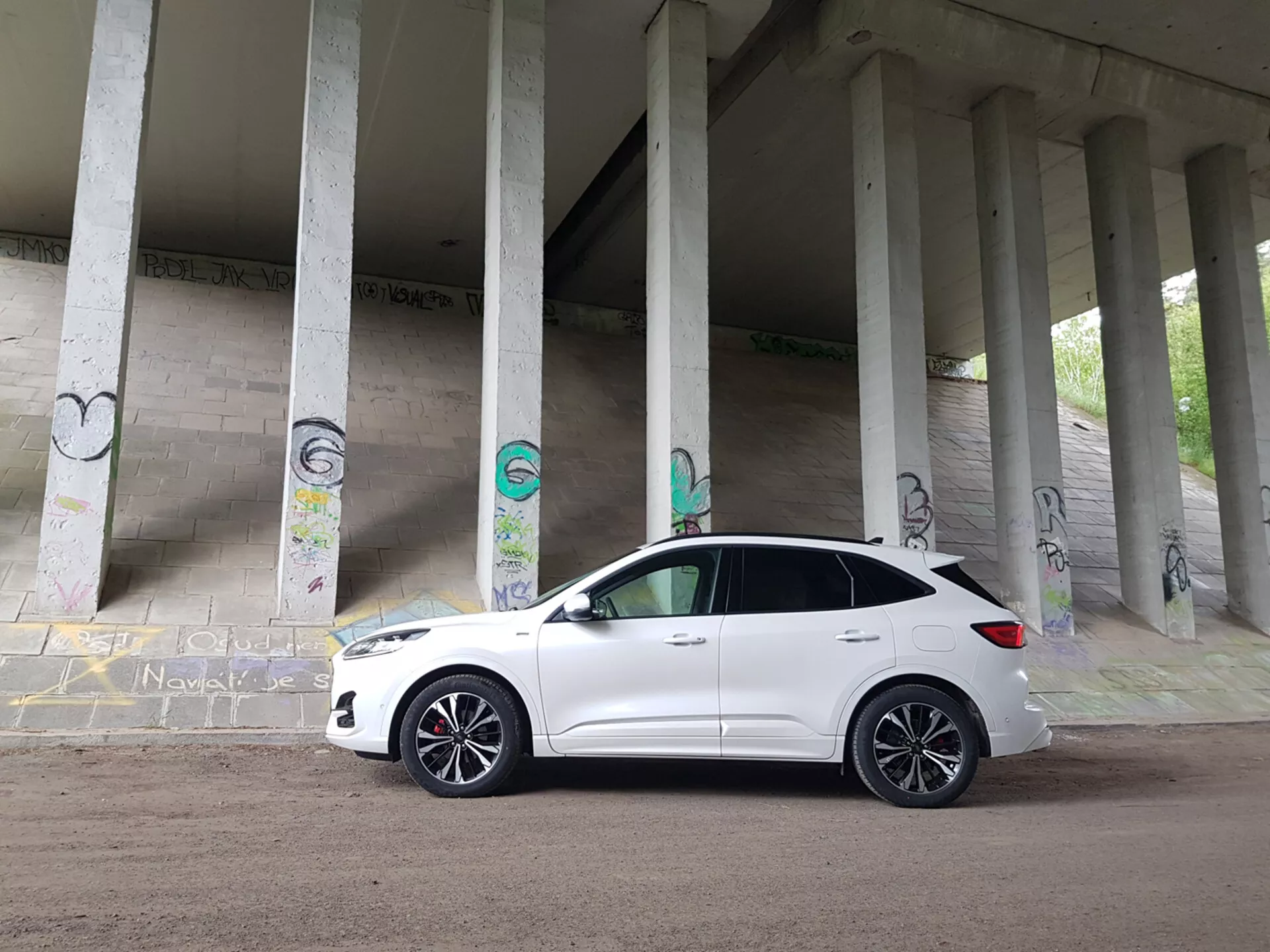 SUV compacto Ford Kuga blanco del 2019 estacionado bajo una estructura con pilares de concreto cubiertos de graffiti, mostrando su perfil frontal incluyendo la parrilla, los faros y las ruedas de aleación