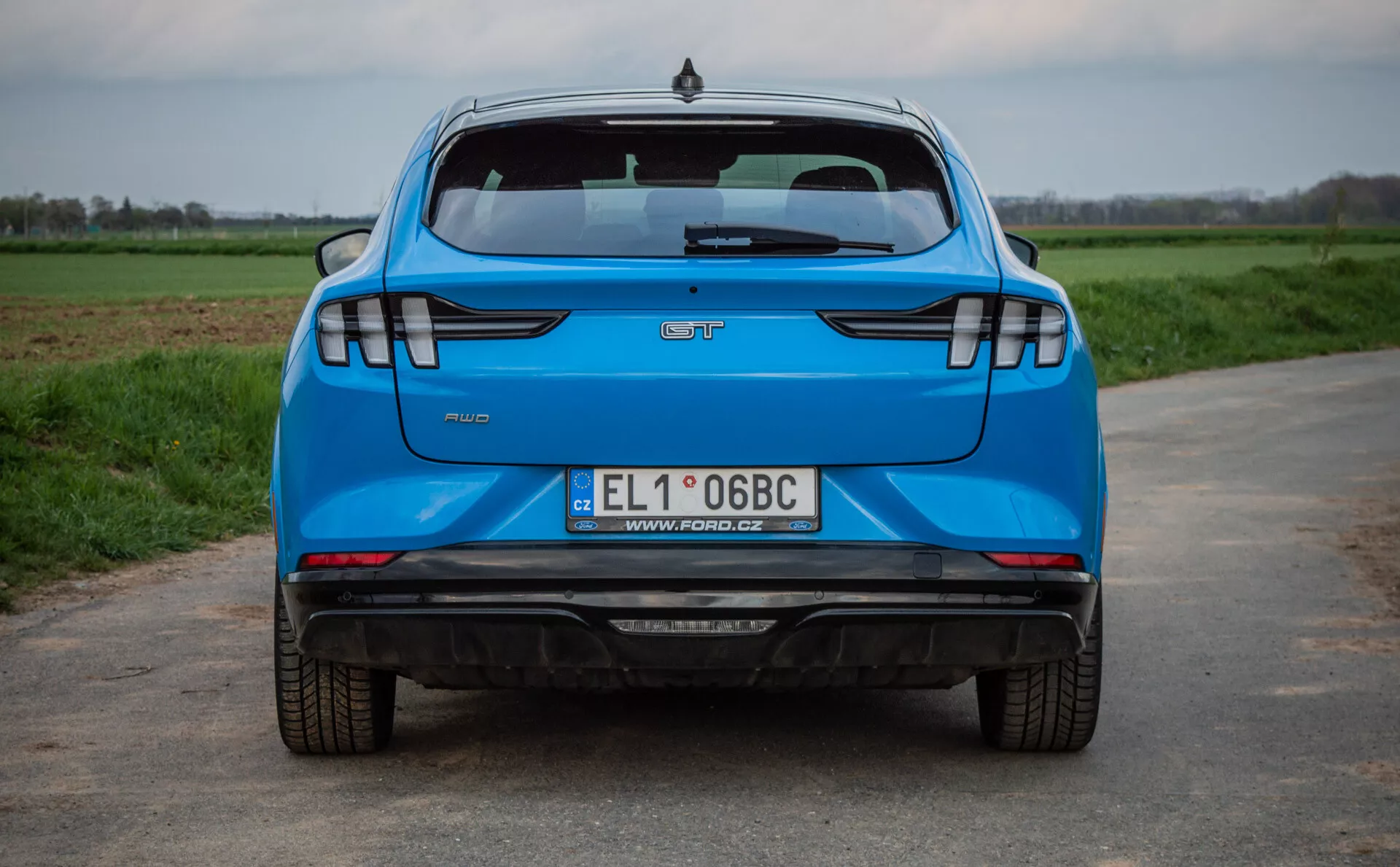 Vista trasera de un Ford Mustang-Mach-E azul del 2020 en una carretera de asfalto con campos de hierba en el fondo, mostrando características clave como el logotipo del fabricante, el nombre del modelo, la insignia del nivel de acabado, la placa de matrícula polaca, sistema de escape doble, luces traseras LED modernas, sensores de estacionamiento y una luz de freno alta montada.