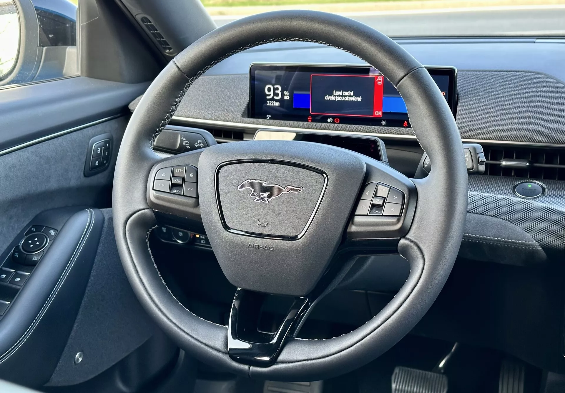 Interior del Ford Mustang Mach-E 2020 destacando el volante moderno de tres radios con botones multifunción y el cuadro de instrumentos digital desde la perspectiva del conductor, mostrando los controles, el emblema y señales de navegación.
