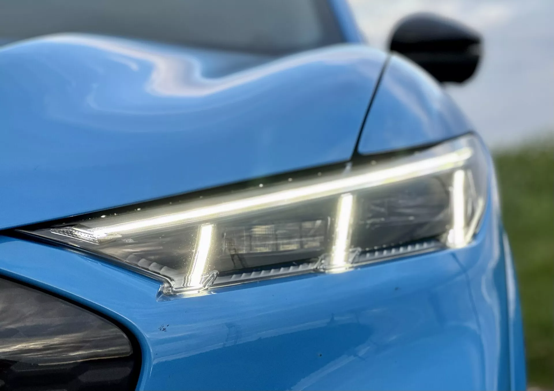 Vista detallada de la sección frontal izquierda del Ford Mustang-Mach-E 2020, destacando el elegante faro LED, el vibrante color azul, y las líneas dinámicas de la carrocería, con un vistazo al espejo lateral y el parachoques delantero, mostrando el moderno diseño y estética.