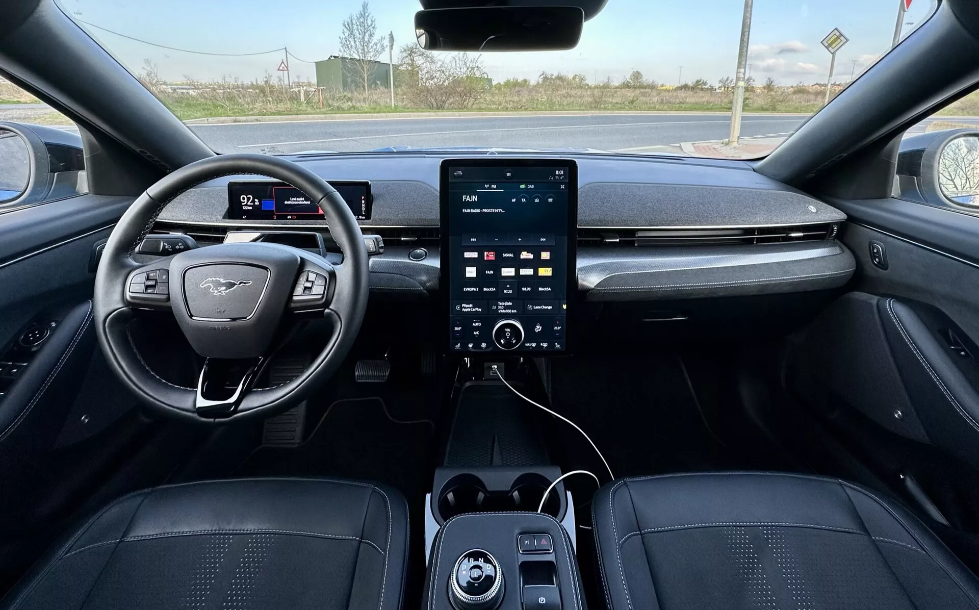 Vista en primera persona del interior del Ford Mustang Mach-E 2020, mostrando el volante posicionado a la izquierda con diseño de fondo plano y emblema, cuadro de instrumentos digital, sistema de infotenimiento con pantalla táctil vertical grande en la consola central con botones de control climático abajo, puertos USB, y tapicería lujosa en tonos oscuros con costuras detalladas, insinuando características avanzadas de seguridad y asistencia, con un fondo exterior rural visible a través del parabrisas.