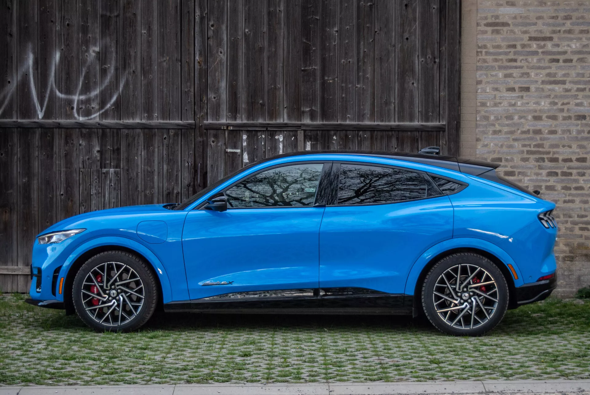 SUV crossover azul Ford Mustang Mach-E 2020 aparcado paralelamente a una puerta de garaje de madera rústica con graffiti, vista lateral que destaca el diseño aerodinámico, línea de techo inclinada estilo coupé, modelo de cuatro puertas con manijas integradas, espejos laterales aerodinámicamente estilizados con señales de giro, ruedas de aleación grandes en dos tonos con pinzas de freno rojas, altura moderada del suelo, carrocería contorneada con líneas esculpidas y pasos de rueda pronunciados, línea de ventanas estrecha con pequeña ventana cuarta después del pilar C, y faldones laterales deportivos