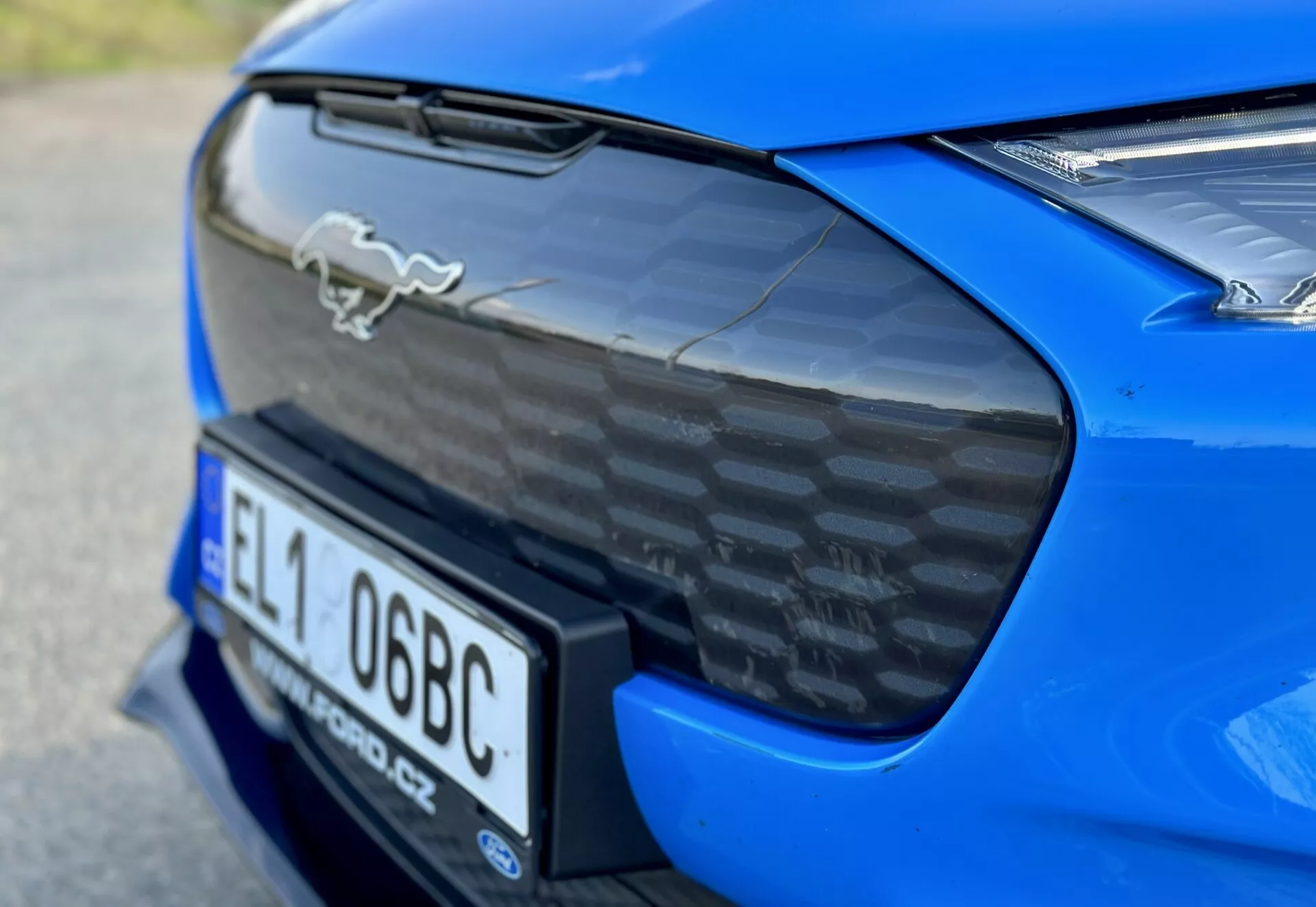 Primer plano de la sección trasera de un Ford Mustang Mach-E 2020 azul, mostrando el emblema de caballo cromado, spoiler trasero de fibra de carbono, nicho integrado para placa de matrícula trasera y vista parcial de luces traseras LED, acentuando las características aerodinámicas y de rendimiento del coche.