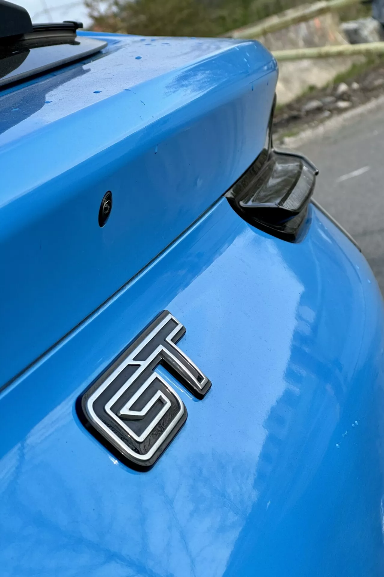 Detalle de la sección trasera de un Ford Mustang Mach-E azul del 2020 con enfoque en la insignia metálica GT, el maletero curvado y el spoiler trasero aerodinámico con un tapón de extremo negro, contra un fondo suavemente desenfocado.