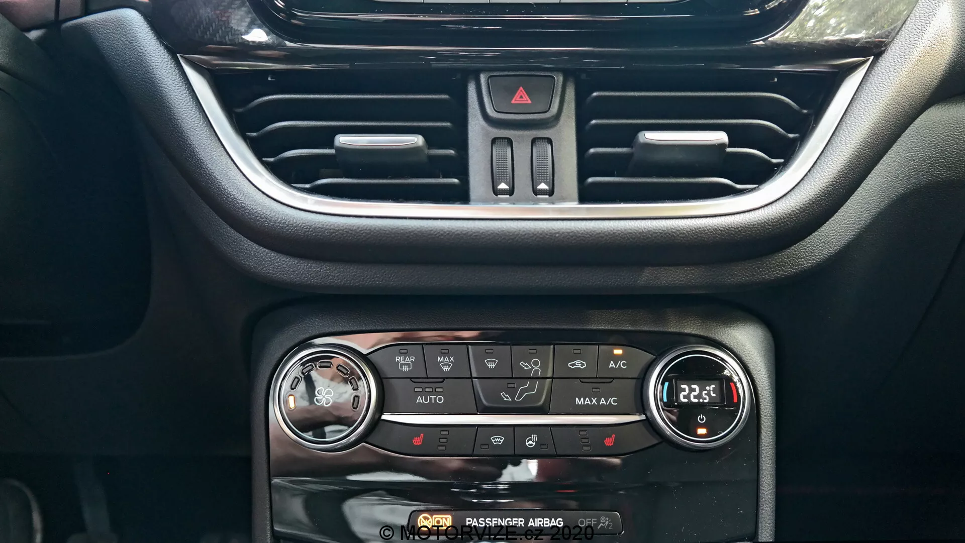 La imagen captura la consola central del tablero del Ford Puma 2020, destacando el sistema de control climático y parte del sistema de infoentretenimiento. Desde un ángulo frontal, la rejilla de ventilación central con láminas horizontales ajustables es visible en la parte superior. Debajo se encuentra el botón triangular rojo de las luces de emergencia. El panel de control climático muestra una variedad de botones y dos diales para ajustar la temperatura y el flujo de aire, con funciones de control climático AUTO, A/C, control de zona dual, desempañado trasero y recirculación. Cada dial de temperatura tiene una pantalla digital, configurada a 22,5 grados Celsius. En el borde inferior está presente el indicador 'AIRBAG PASAJERO DESACTIVADO'. El diseño interior integra plásticos negros modernos y acentos plateados para una interfaz amigable.