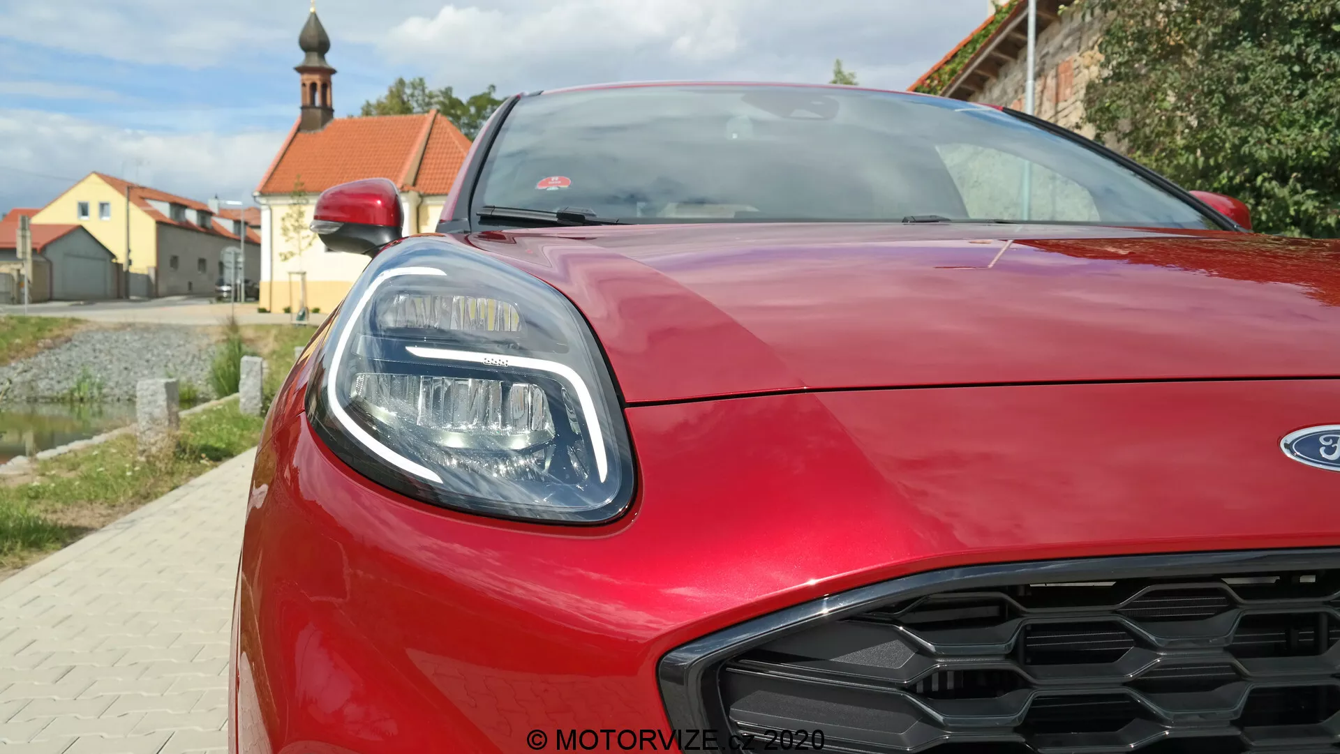 Vista frontal en primer plano de un Ford Puma rojo 2020, destacando el faro derecho con elementos LED elegantes, capó aerodinámico esculpido y diseño de rejilla en forma de panal. Imagen tomada desde un ángulo bajo, capturando el estilo moderno y los detalles deportivos del coche.