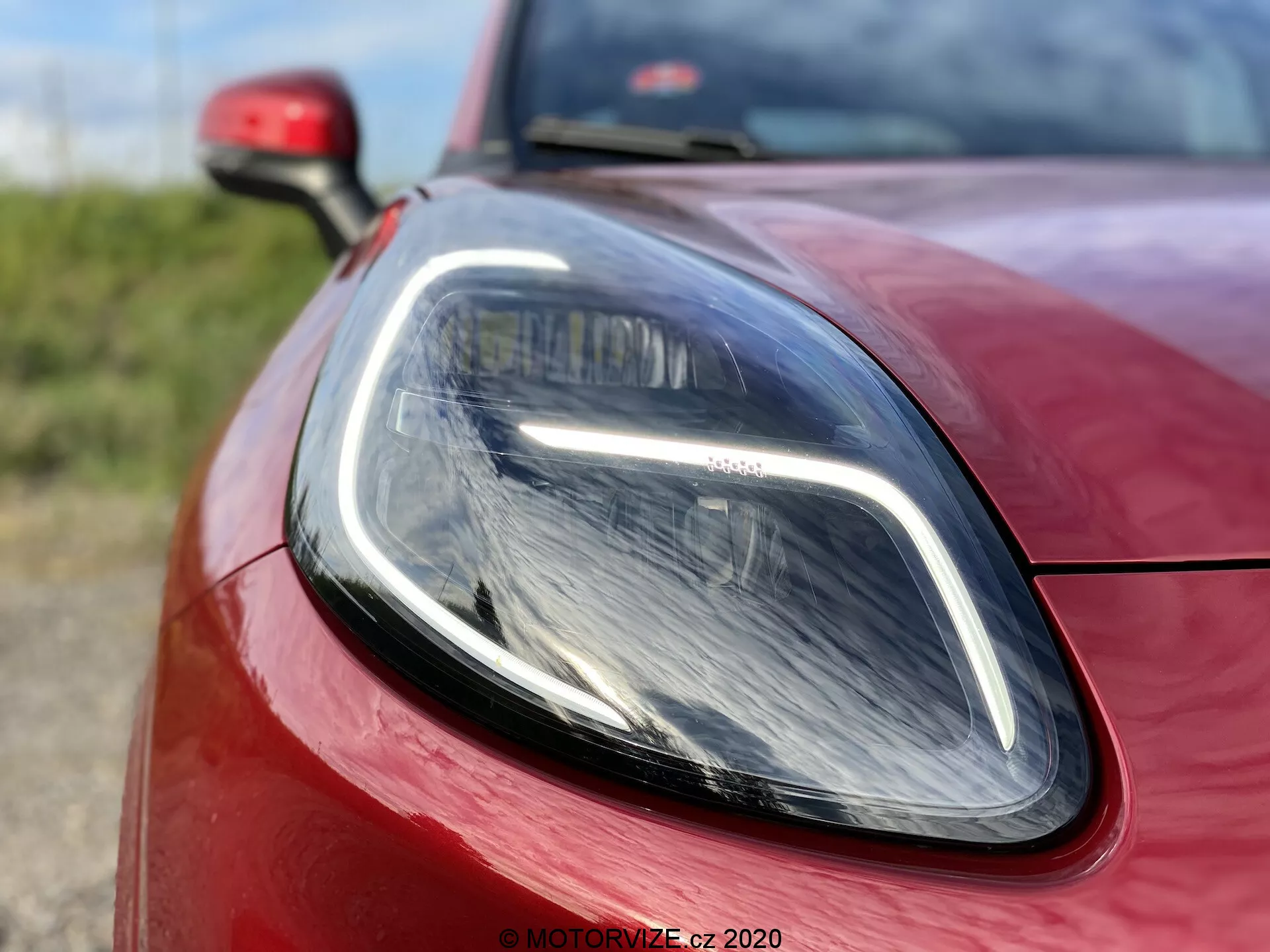 Primer plano del Ford Puma 2020, enfocado en la sección del faro delantero del lado del conductor y la carrocería circundante. Tomada desde un ángulo bajo y ligeramente descentrado, la imagen resalta las luces LED diurnas elegantes y el faro de tipo proyector moderno. La pintura roja vibrante y las alineaciones precisas de los paneles enfatizan el diseño deportivo y aerodinámico del coche.