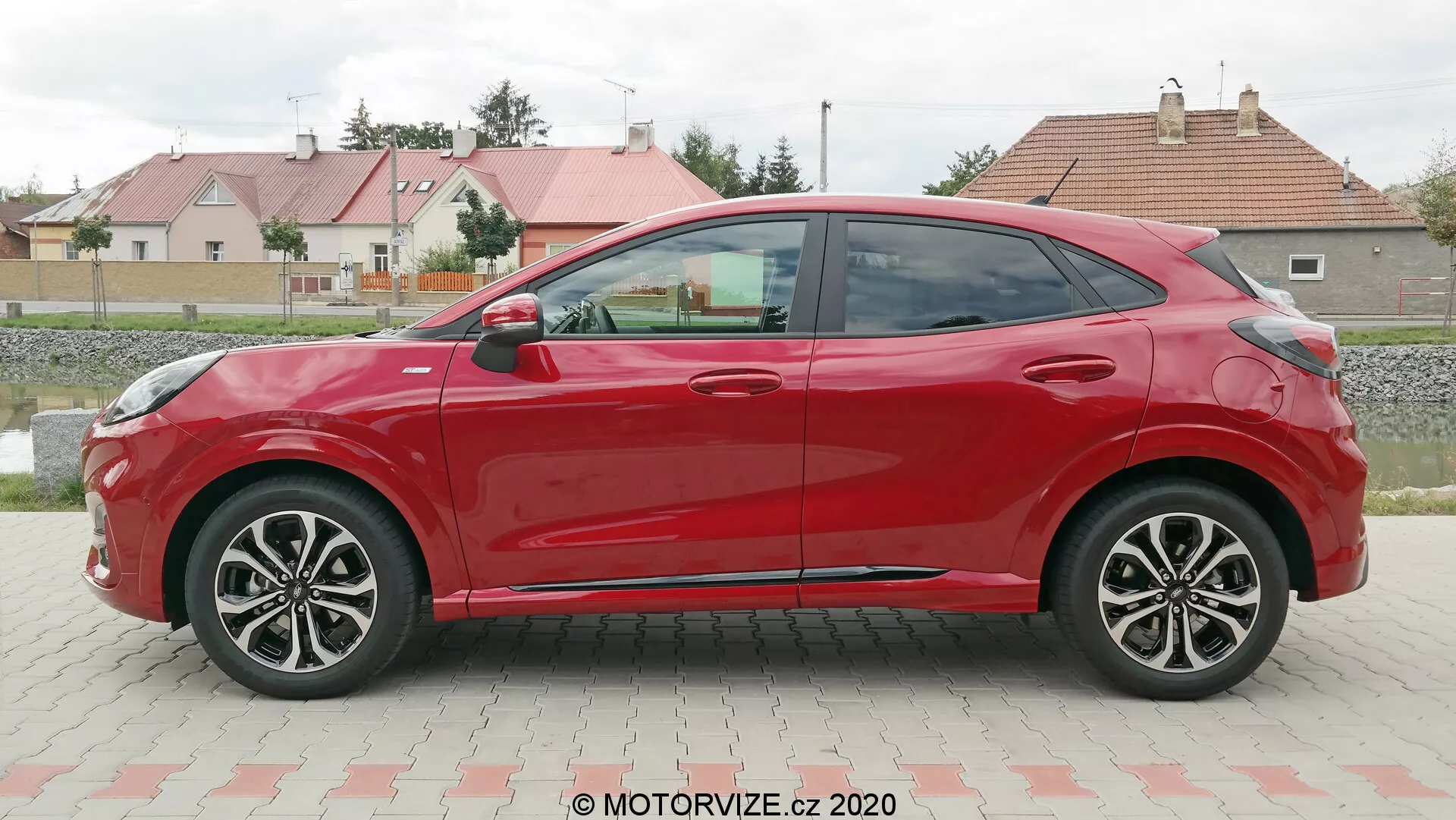 Vista de perfil lateral de un Ford Puma hatchback rojo de cuatro puertas 2020, tomada durante el día en un estacionamiento con un fondo de césped y edificios residenciales. El coche presenta líneas aerodinámicas, llantas de aleación de múltiples radios, espejos y manijas de puertas a juego con el color, y una línea del techo descendente. La ausencia de cerraduras visibles sugiere un posible sistema de entrada sin llave.