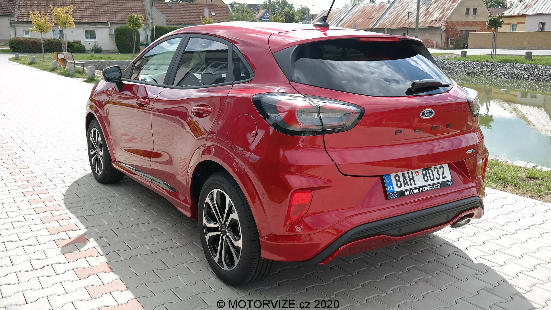 Vista trasera de tres cuartos de un hatchback Ford Puma 2020 rojo vibrante capturada con luz natural diurna. La imagen resalta los contornos aerodinámicos del coche, las distintivas luces traseras que envuelven los lados y los sensores integrados en el parachoques trasero, lo que sugiere capacidades de asistencia de estacionamiento. Los tubos de escape dobles y el alerón trasero aumentan su atractivo deportivo, mientras que las llantas de aleación multirradio y elementos del mismo color de la carrocería, como los espejos laterales y las manijas de las puertas, contribuyen a su diseño moderno. Limpiaparabrisas trasero visible, indicando practicidad en condiciones climáticas adversas.