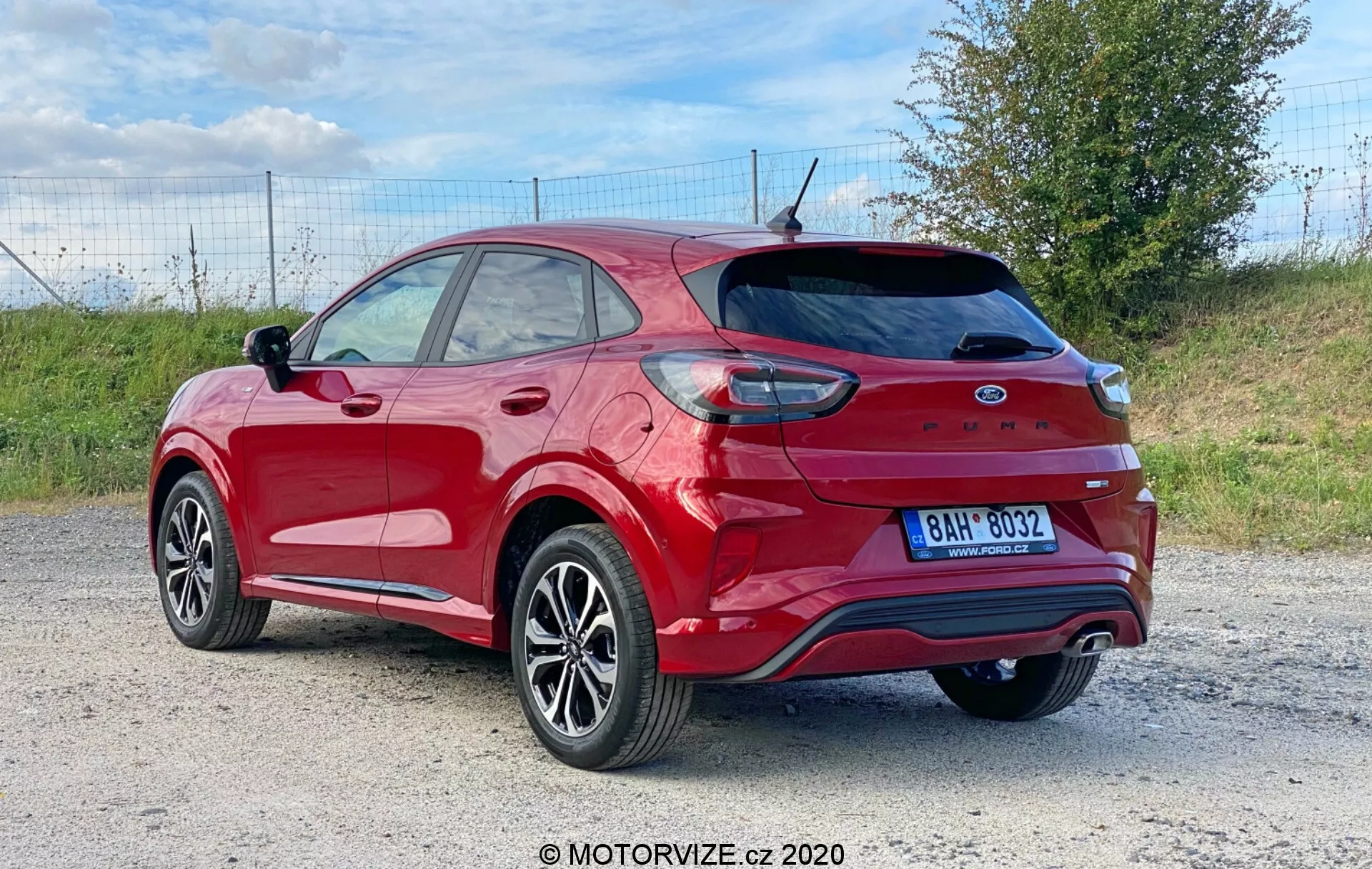 La imagen presenta un Ford Puma 2020 crossover/SUV rojo fotografiado desde un ángulo de tres cuartos trasero, mostrando la rueda trasera y el neumático, parte de la puerta delantera y la puerta trasera de pasajeros, el panel trasero y toda la puerta trasera con spoiler integrado y limpiaparabrisas trasero. Los elementos visibles incluyen luces traseras que envuelven parcialmente el guardabarros trasero, parachoques trasero con reflectores y sensores de aparcamiento, sistema de escape dual, antena de aleta de tiburón, parabrisas trasero con líneas de desescarcha, ruedas de aleación con diseño multi-spoke y neumáticos de perfil bajo, ventanas tintadas, rieles de techo y carcasa de espejo lateral a juego con el color de la carrocería. El vehículo está situado en un entorno al aire libre, posiblemente en una carretera de tierra o de grava, con un tramo de césped en el fondo que sugiere un entorno rural o semi-rural.