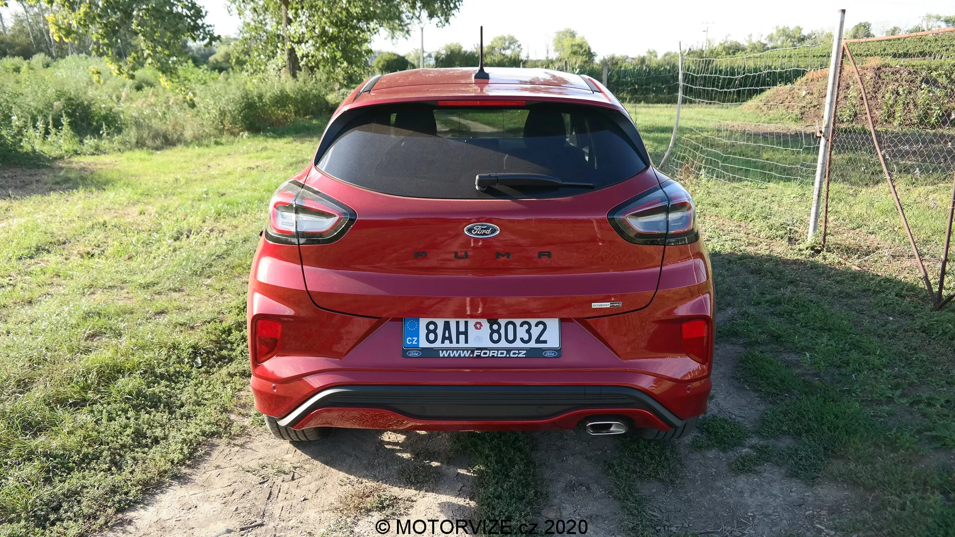 Vista trasera de un Ford Escape 2020 rojo con tecnología híbrida estacionado en una superficie sin pavimentar. El vehículo cuenta con luces traseras modernas, un logo de Ford centrado y una matrícula europea. El parabrisas trasero tiene un limpiaparabrisas y una tercera luz de freno LED. La parte inferior de la carrocería y el parachoques tienen un recubrimiento protector de plástico negro con sensores visibles que indican tecnología de asistencia de estacionamiento. Vegetación en el fondo sugiere un entorno rural.