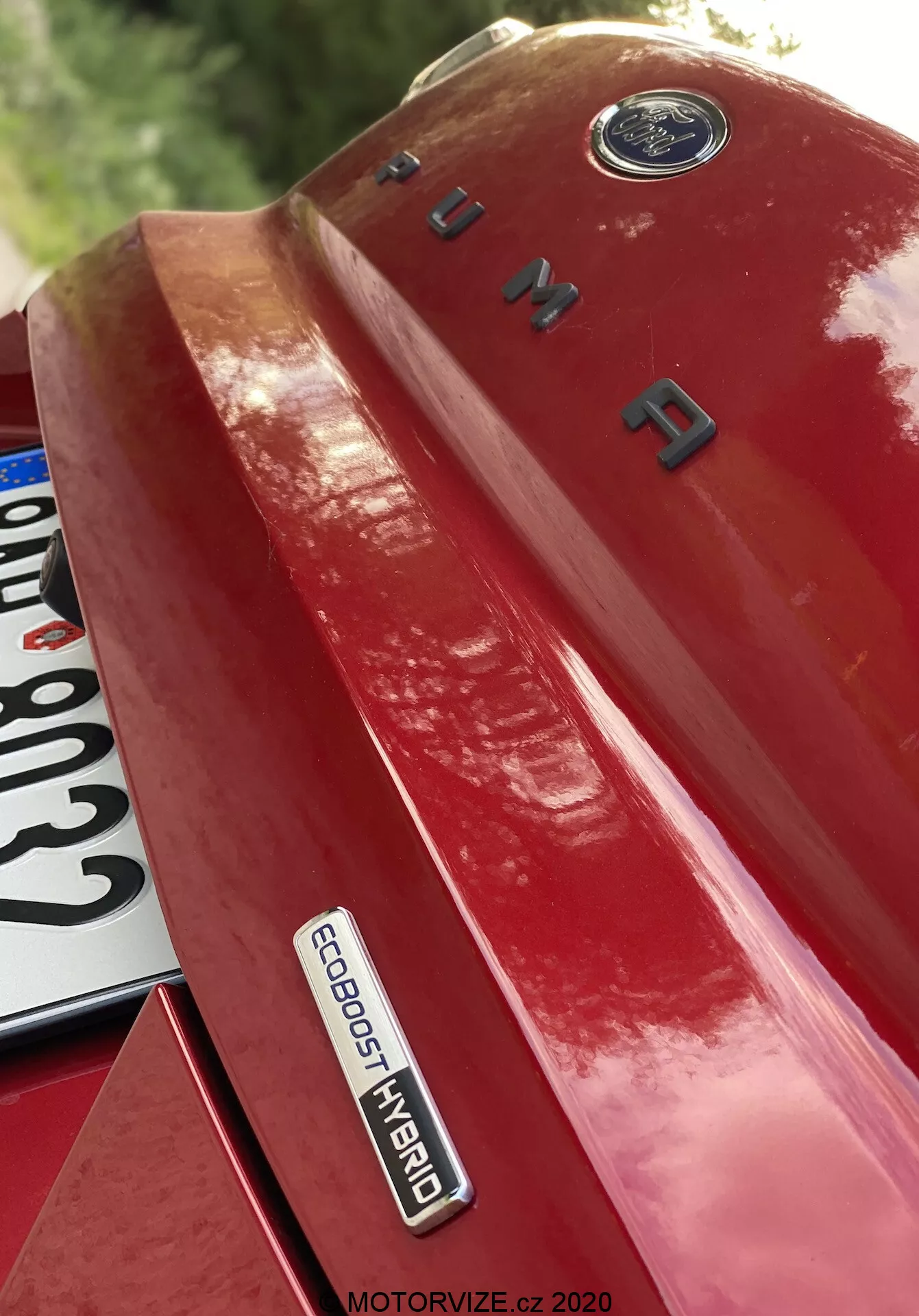Detalle en primer plano del cuarto trasero izquierdo de un Ford Puma 2020 rojo, tomado desde un ángulo ascendente, presentando prominentemente las insignias y la marca. La imagen resalta el emblema de Ford en la parte superior, el nombre del modelo 'PUMA' en letras cromadas en el maletero y la insignia 'ECOBOOST HYBRID', que indica una avanzada tecnología híbrida. Una porción de la matrícula es parcialmente visible en la parte inferior izquierda. La pintura brillante refleja la luz, acentuando el diseño y la sofisticación tecnológica del vehículo.